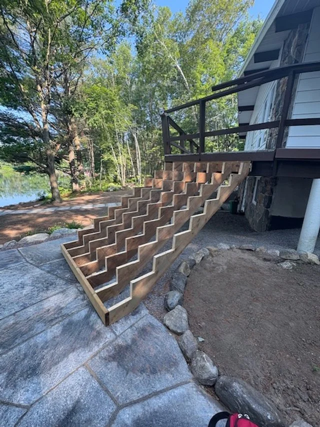 Wooden staircase leading from a patio to a deck on a house, with a landscape of trees and a body of water in the background.