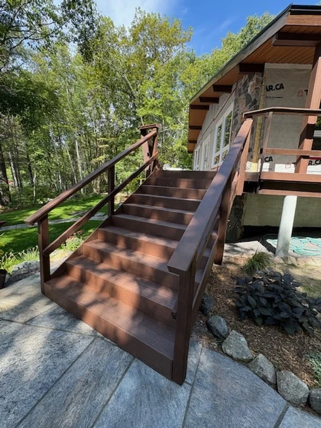 Wooden exterior staircase with handrails leading up to a house, surrounded by trees and a landscaped yard.