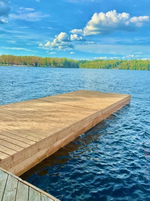 A wooden dock extends over a calm lake, with a tree-lined shoreline and a partly cloudy sky in the background.