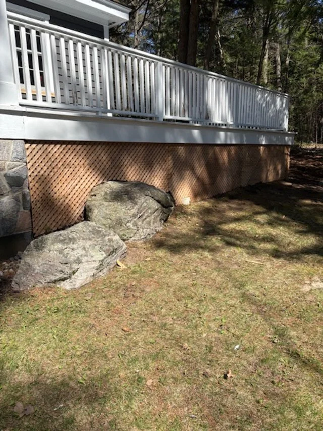 Backyard with a raised deck with white railing, brick skirting, and two large rocks on the ground below in a grassy yard with trees in the background.