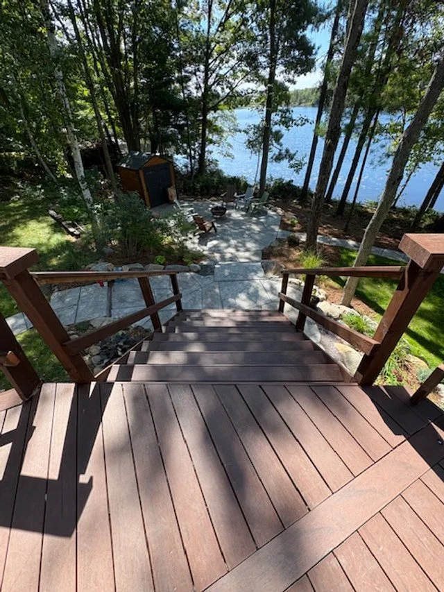 A view from the top of a wooden staircase leading down to a lakeside yard with trees, outdoor furniture, and a boat house on the water.