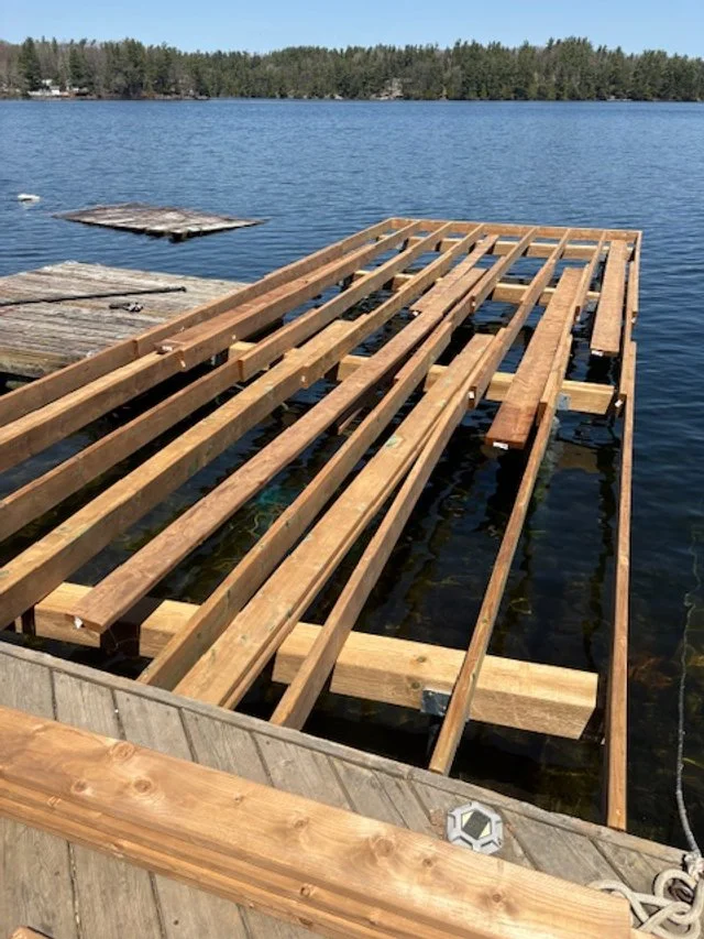Under construction wooden dock extending into a calm lake, with trees in the background.