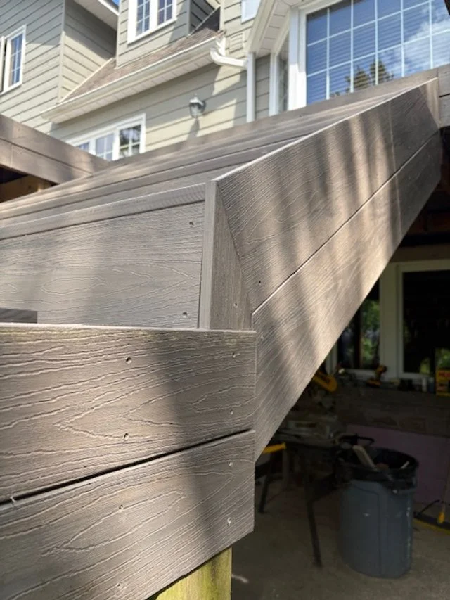 Close-up of a composite deck support beam showing detailed wood grain with a house and window in the background.