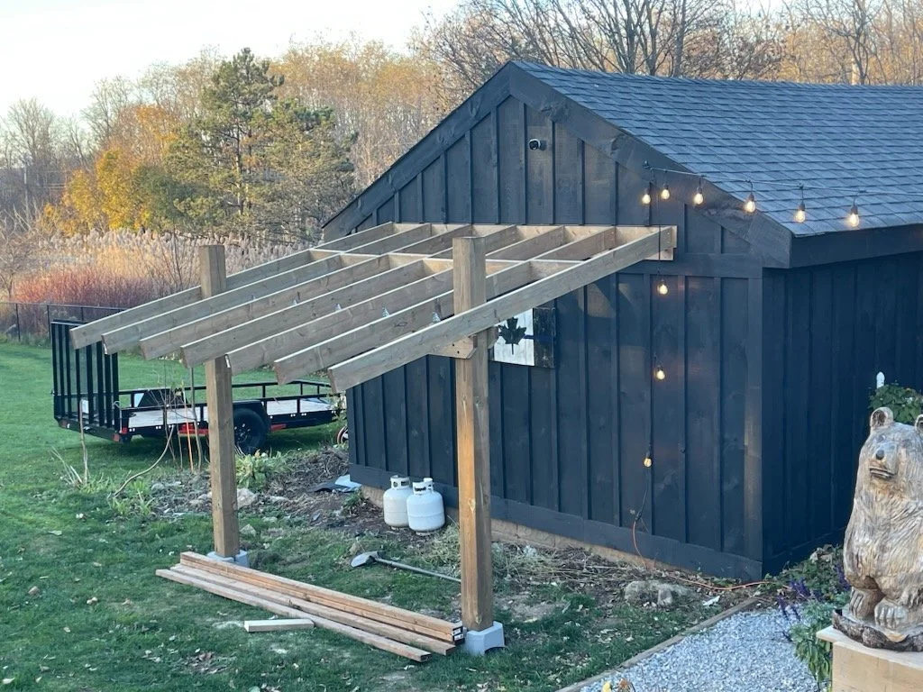 Wooden frame structure under construction next to a dark blue shed, with string lights hanging, and a trailer parked nearby in a yard with trees in the background.