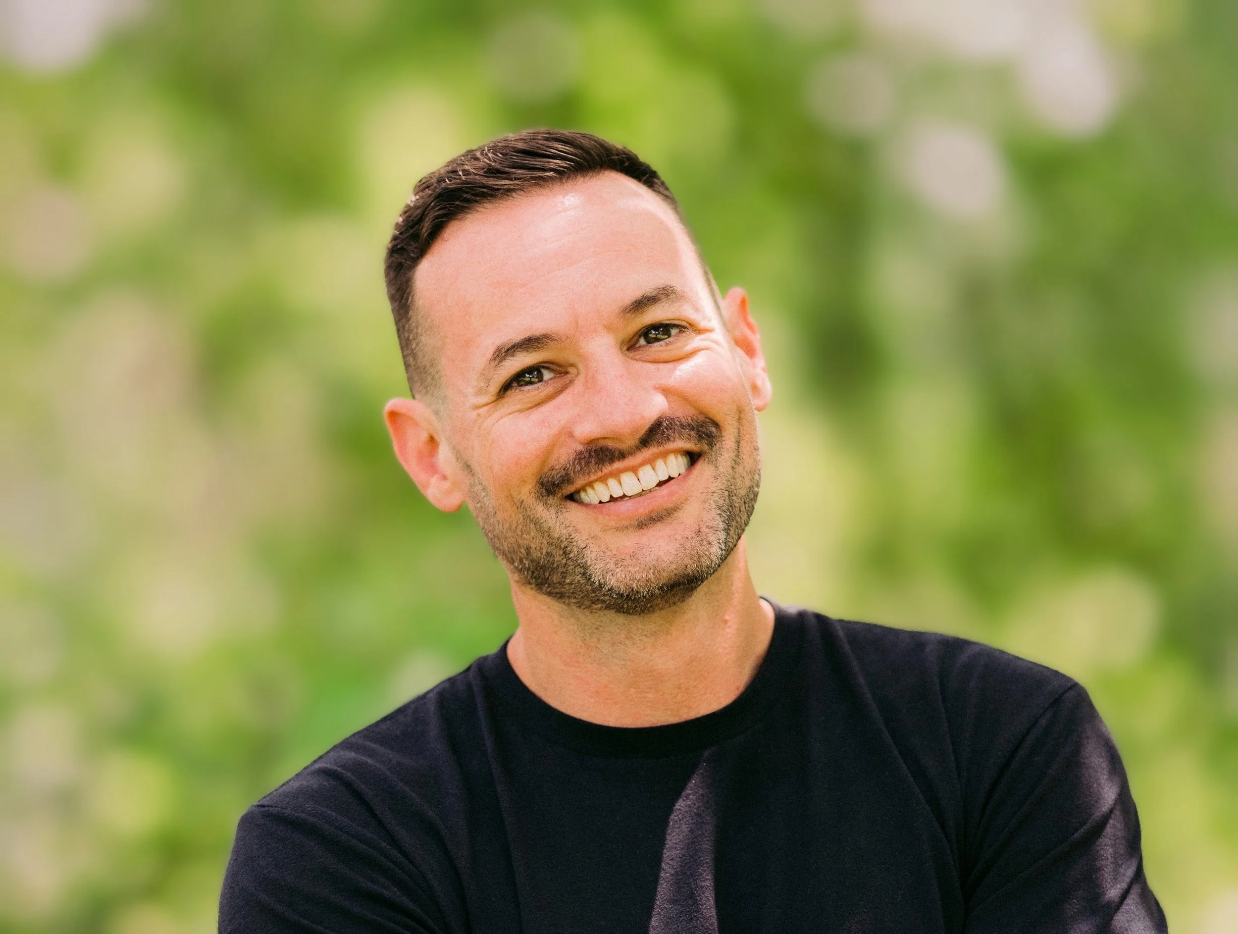 A smiling man with short dark hair and a beard, wearing a black shirt, outdoors with a blurred green background.