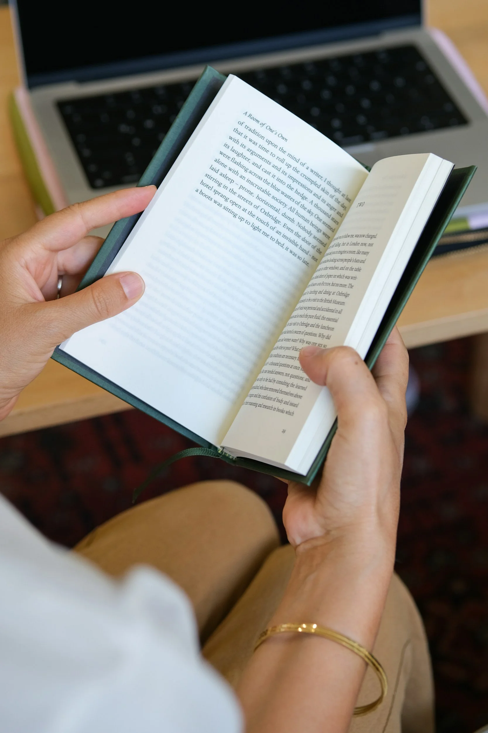 Person holding an open book while sitting at a table with a laptop in the background.