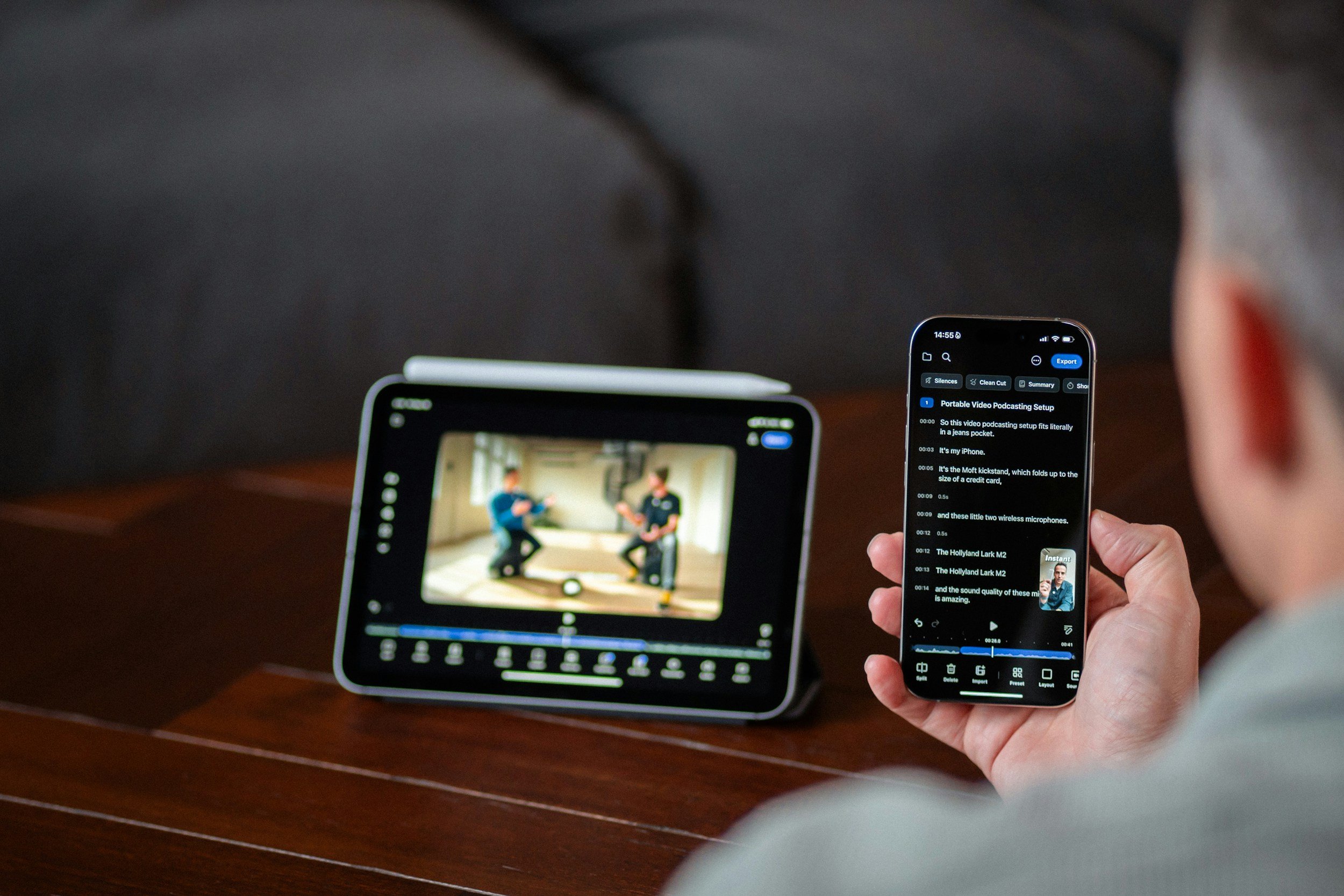 Person holding a smartphone while editing a video or audio file on it, with a small screen showing a video of two people in a room, and the phone displaying a transcript or notes related to the recording.