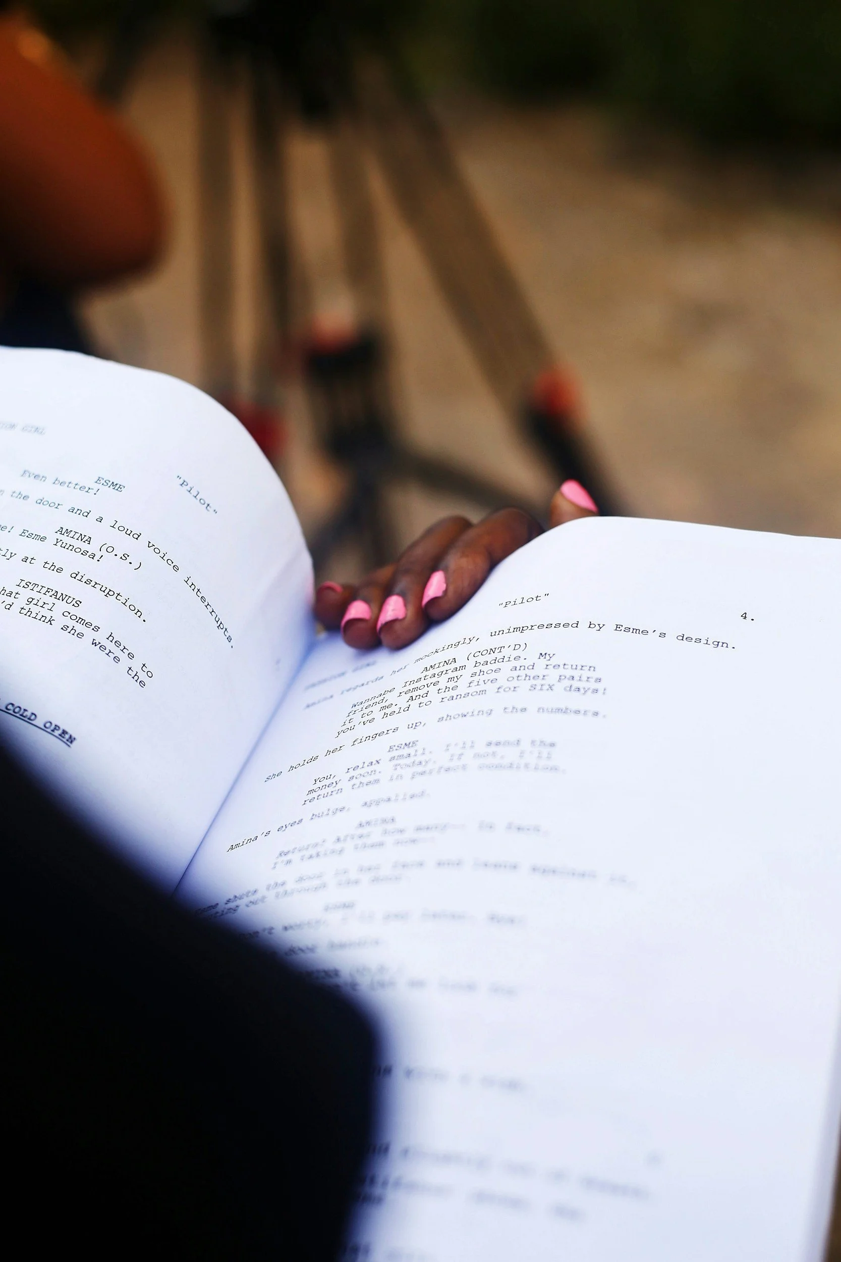 Person with pink painted nails holding a script or screenplay, with a ladder and outdoor setting in the background.