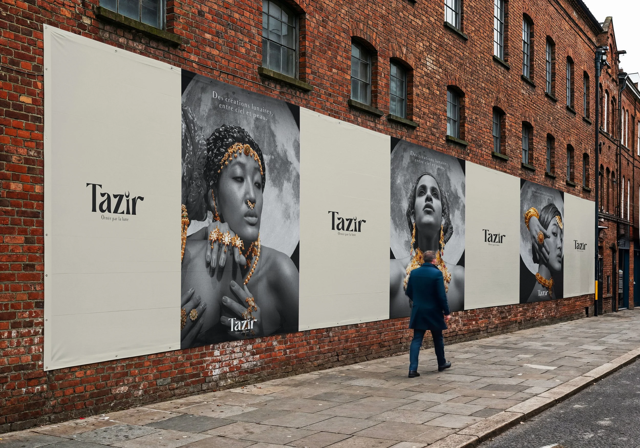 Un homme marche devant une affiche publicitaire en noir et blanc sur un mur de briques rouges. L'affiche montre des portraits de femmes portant des bijoux, avec le texte 'Tazir' et 'Oindre par la lune'.
