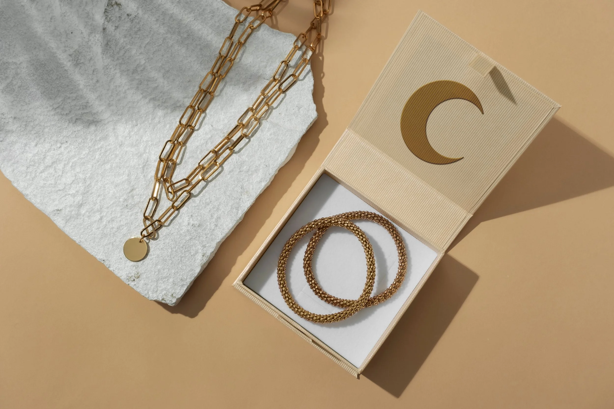 Collier en or, trois bracelets en or dans une boîte en carton avec un motif de croissant de lune, sur fond beige avec une pierre blanche.