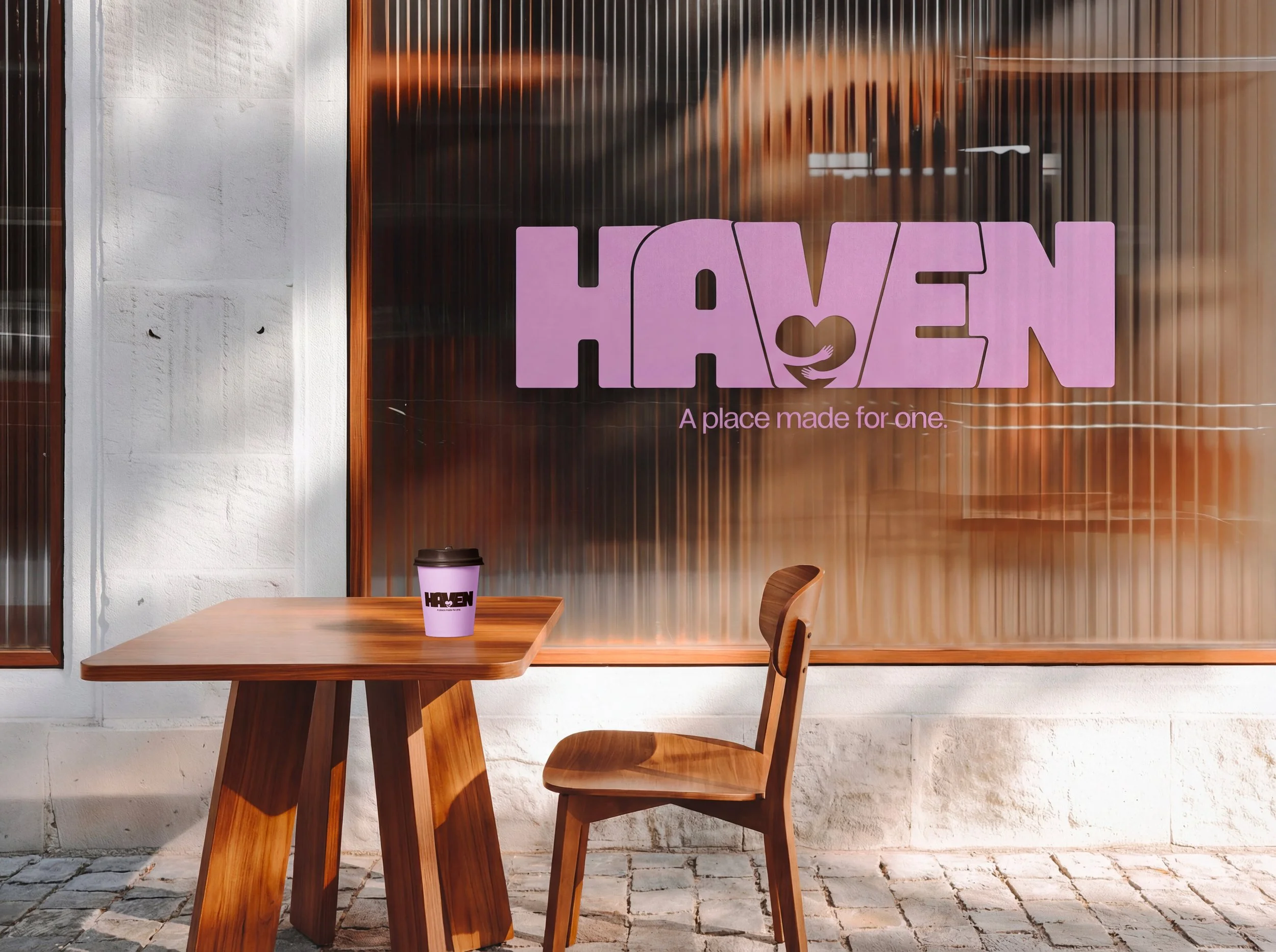 Un meuble en bois avec une tasse à café en papier portant l'inscription HAVEN, placé devant une façade en verre avec un logo violet HAVEN et le slogan "A place made for one".