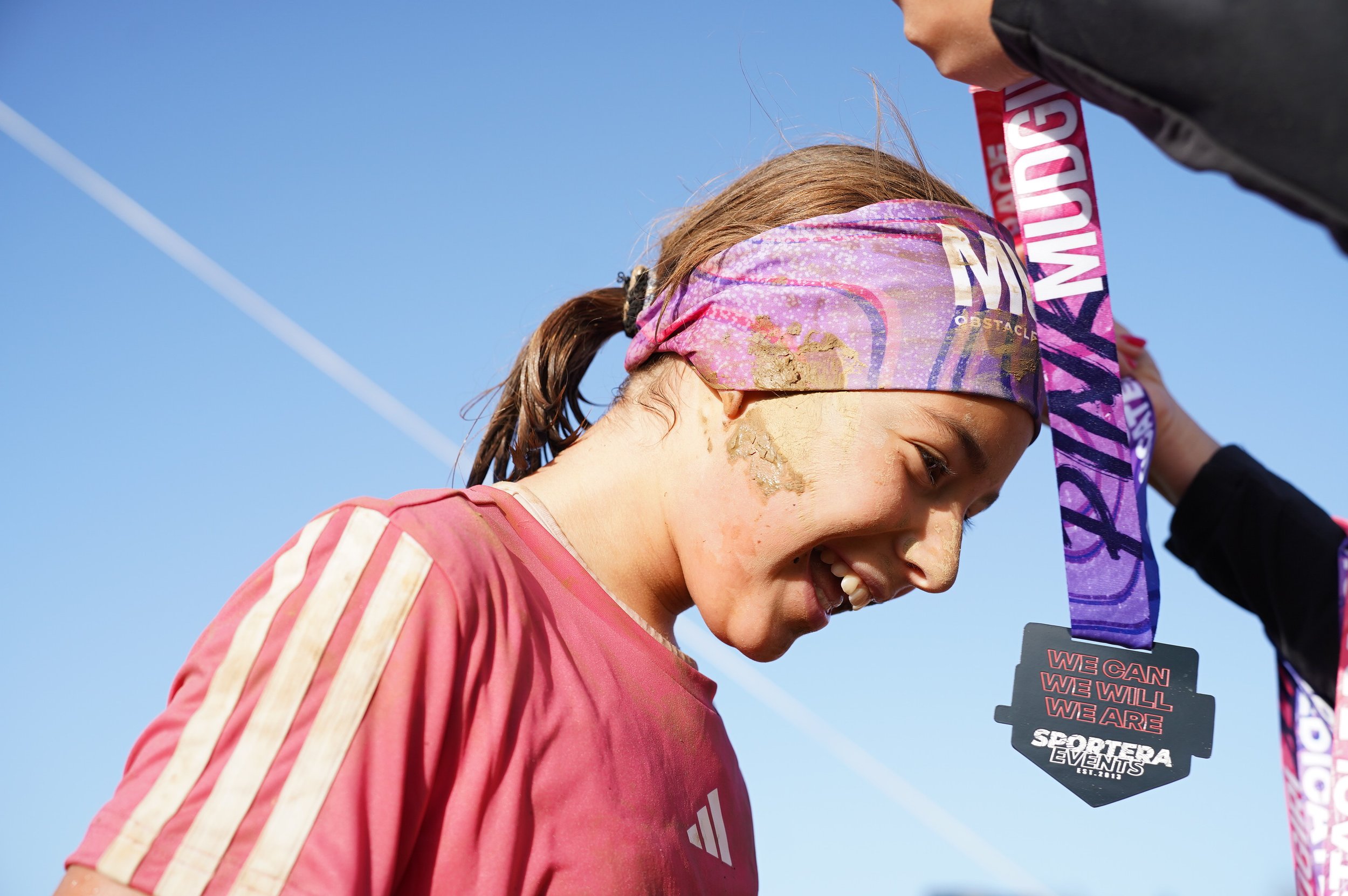 Une jeune fille souriante reçoit une médaille après une compétition, avec le ciel bleu en arrière-plan.