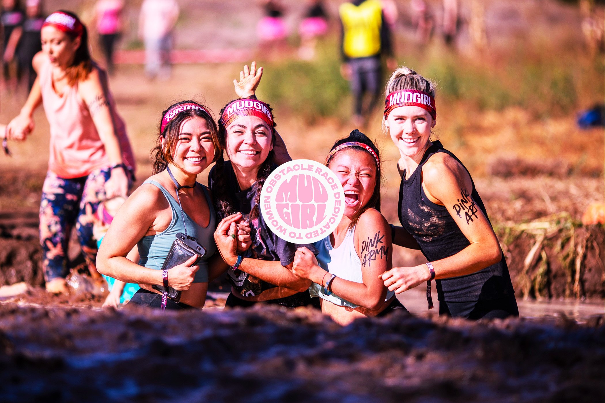 Groupe de femmes souriantes et joyeuses participant à une course d'obstacles à travers la boue, portant des bandeaux roses et des tatouages temporaires. Elles tiennent un panneau indiquant 'Women Obstacle Race Designed to Girl'.