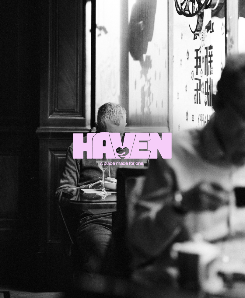 Jeune garçon assis seul à une table dans un restaurant, privant la majorité de son visage. Mots 'Haven' en rose avec le slogan 'A place made for one' en dessous, sur une image en noir et blanc.