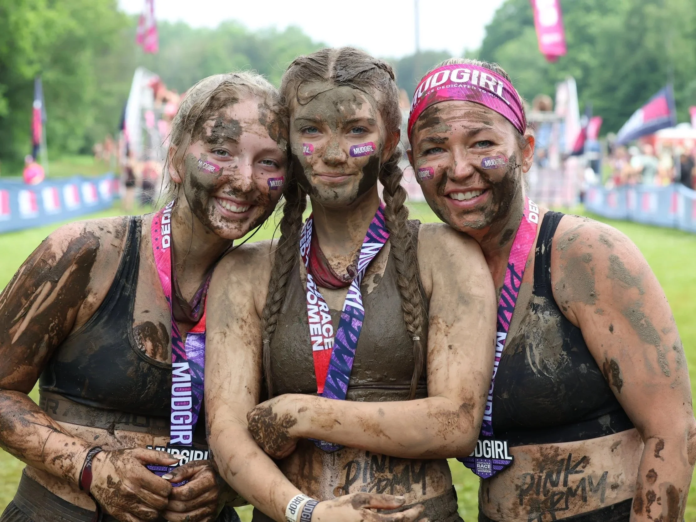 Trois femmes souriantes après une course où elles ont été couvertes de boue, portant des médailles et des bandeaux roses lors d'un événement Muddy Girl Mud Run. L'environnement est une zone extérieure verdoyante avec une foule en arrière-plan.