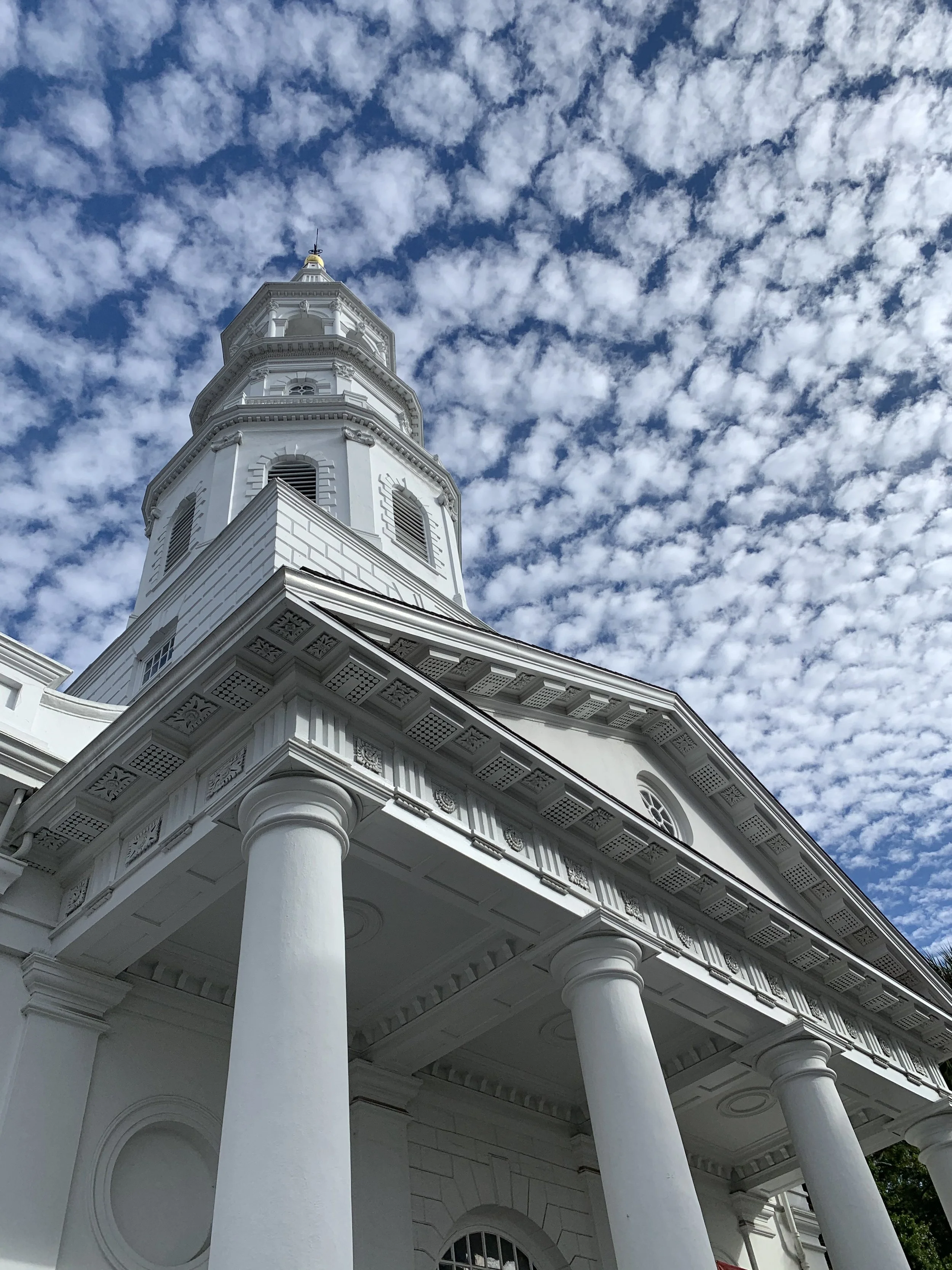 A white historic building with pillars and a tall steeple against a partly cloudy sky.