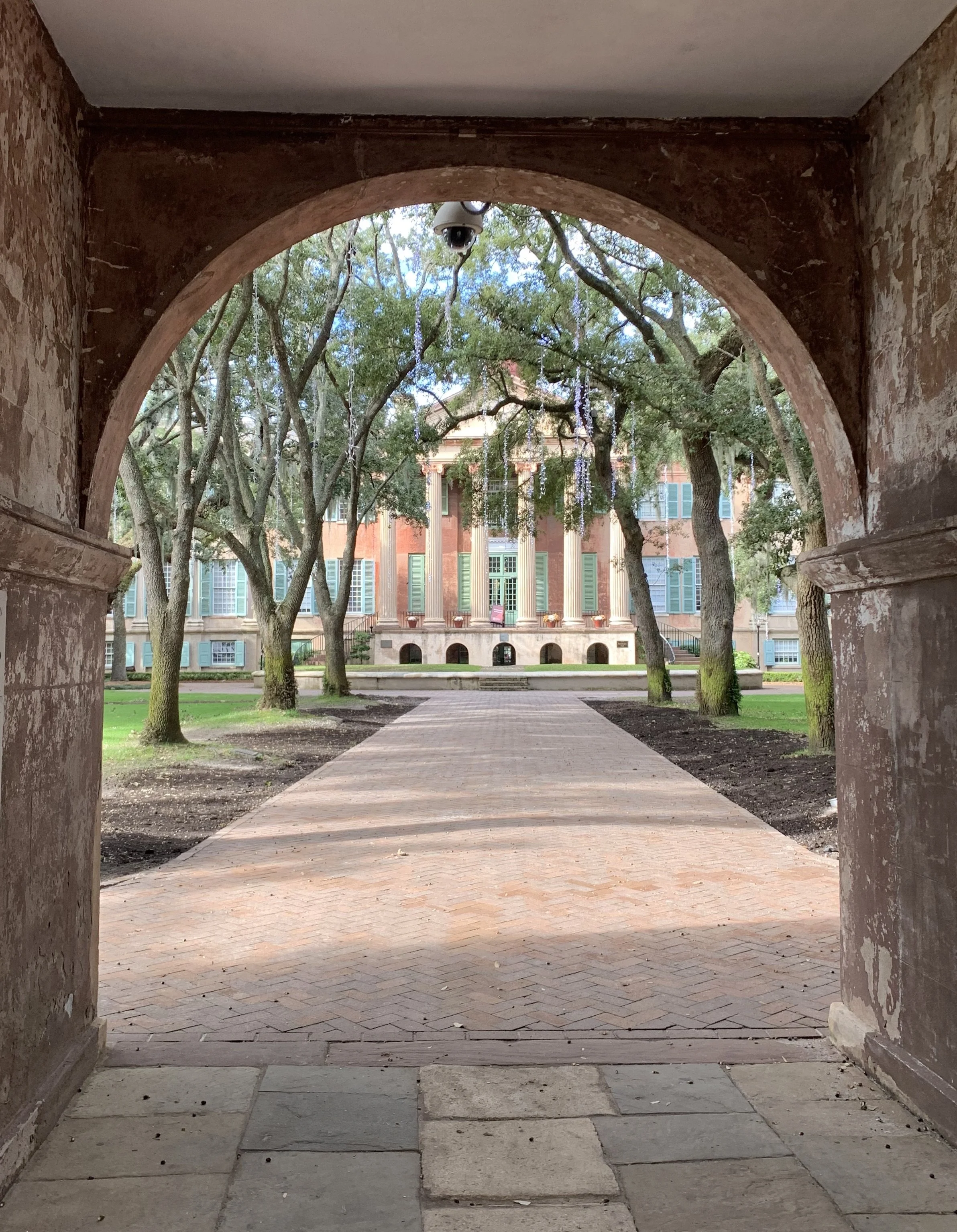 View through an archway of a historic brick building with green shutters, surrounded by trees, and a brick pathway leading to the building's steps.