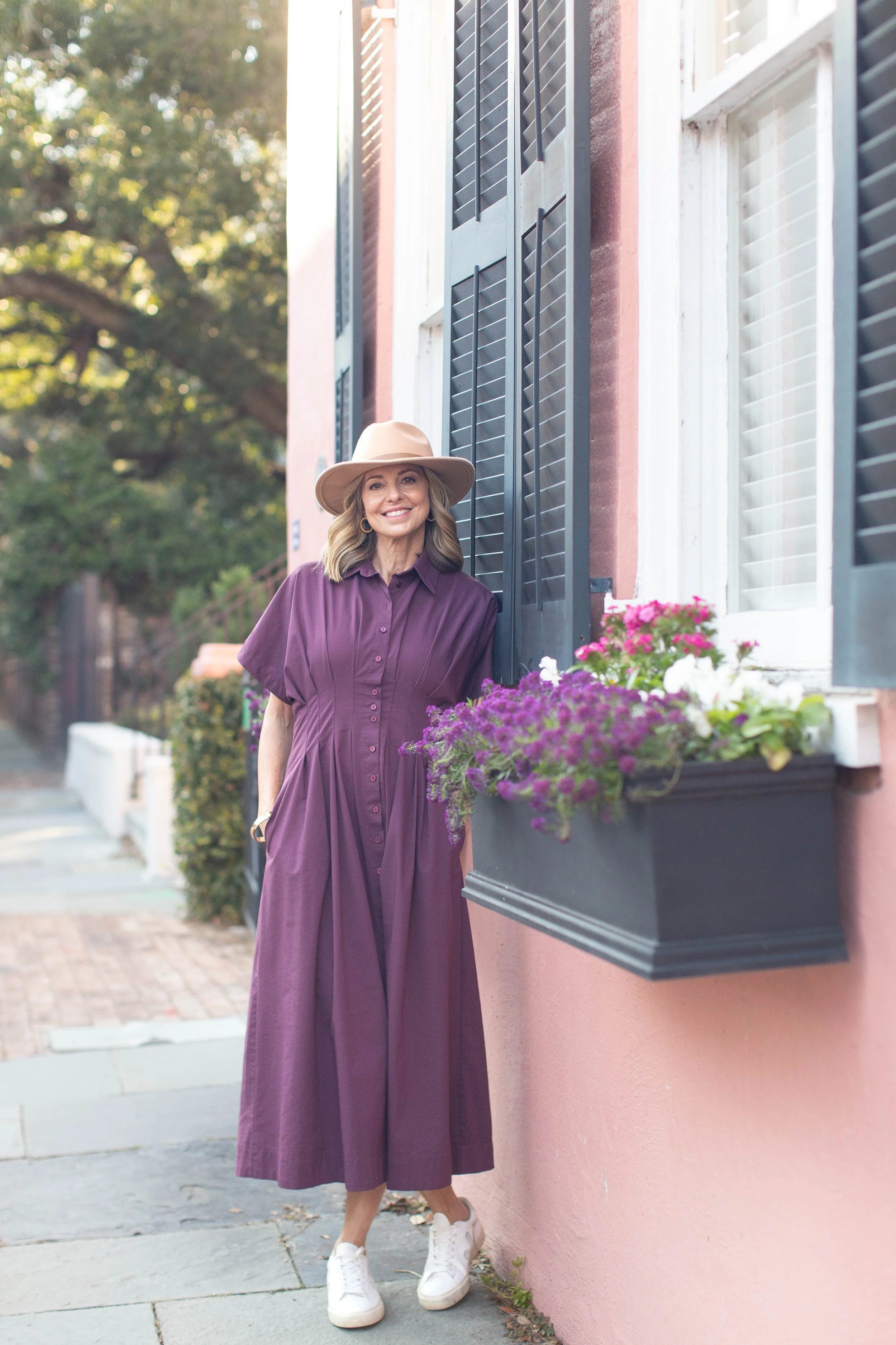 Rachel Fair licensed Charleston tour guide, wearing a wide-brimmed hat and a purple button-up dress with pleats, smiling and standing beside a pink wall with black window shutters and a window box filled with pink, purple, and white flowers.