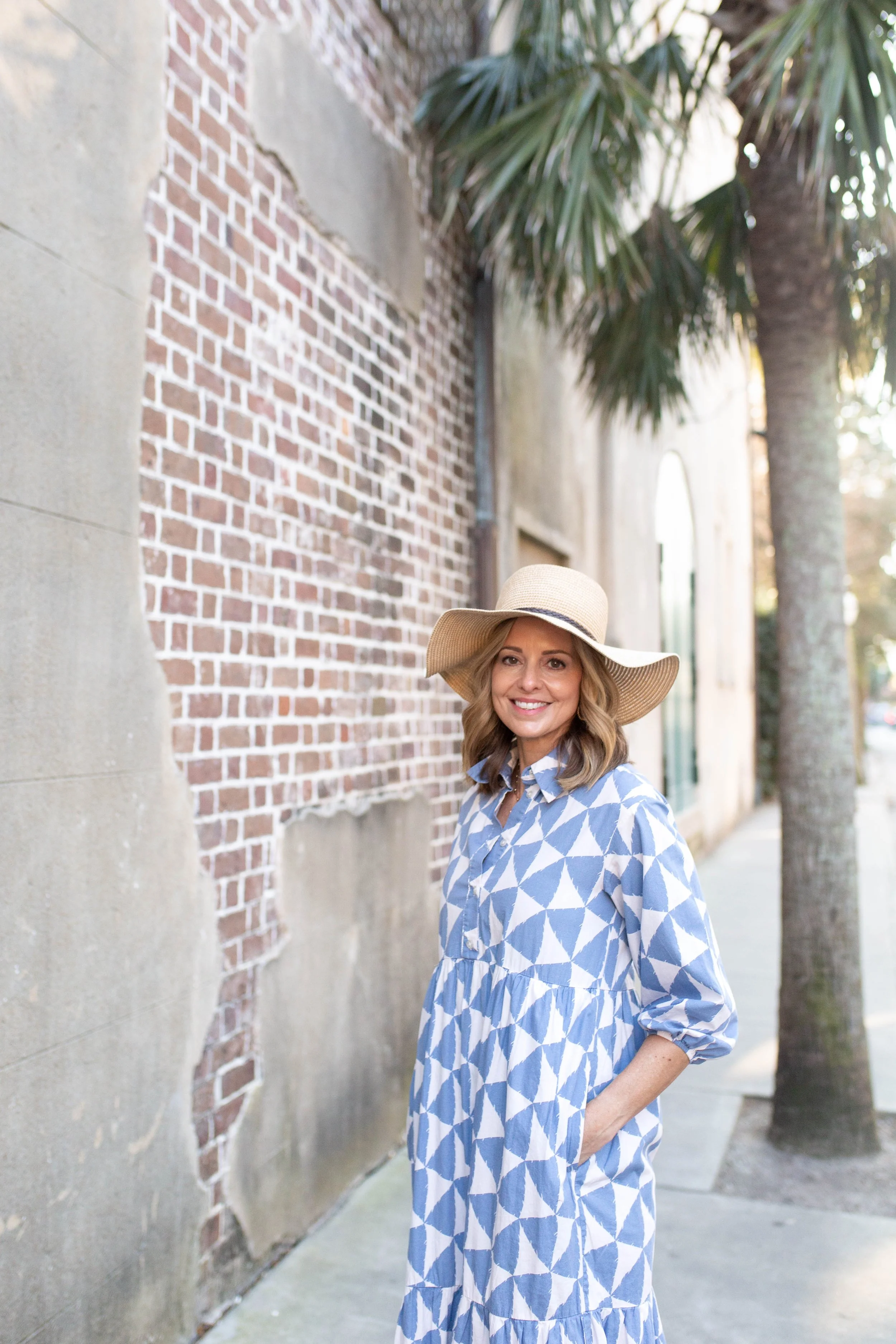 Rachel Fair licensed Charleston tour guide, standing on a sidewalk next to a brick wall with palm trees in the background, wearing a wide-brimmed hat and a blue and white patterned dress.