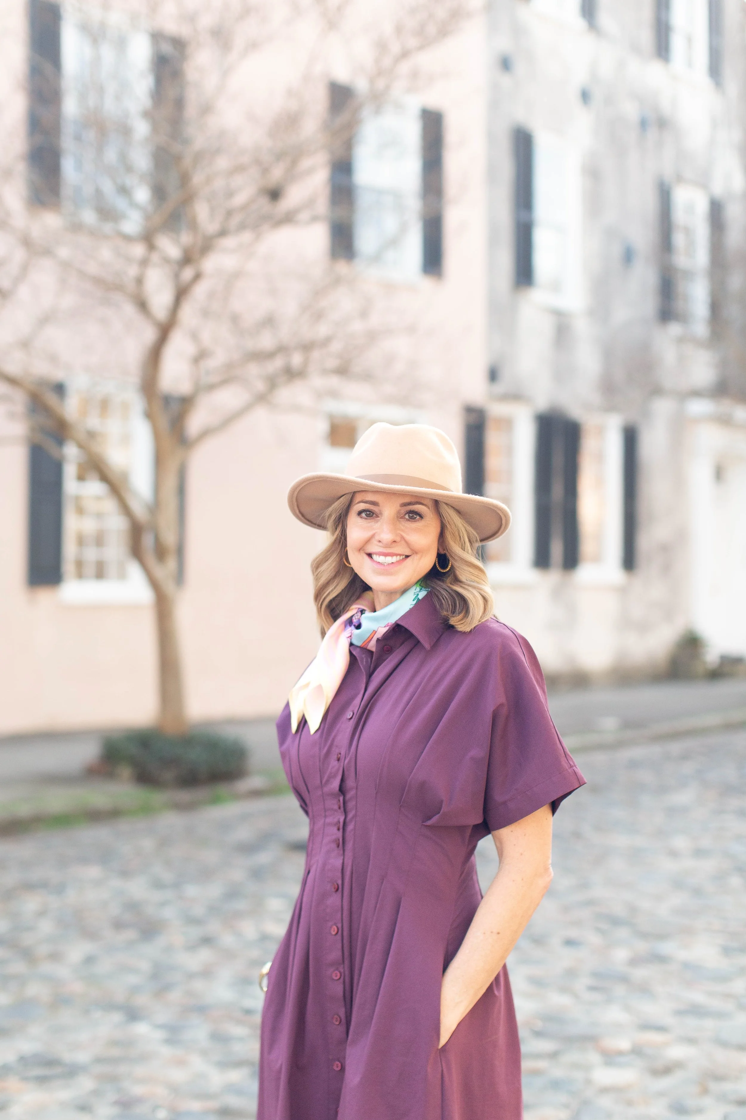 Rachel Fair licensed Charleston tour guide, wearing a beige hat, purple dress, and a light-colored scarf, stands outdoors on a cobblestone street with historical buildings and a leafless tree in the background, smiling at the camera.