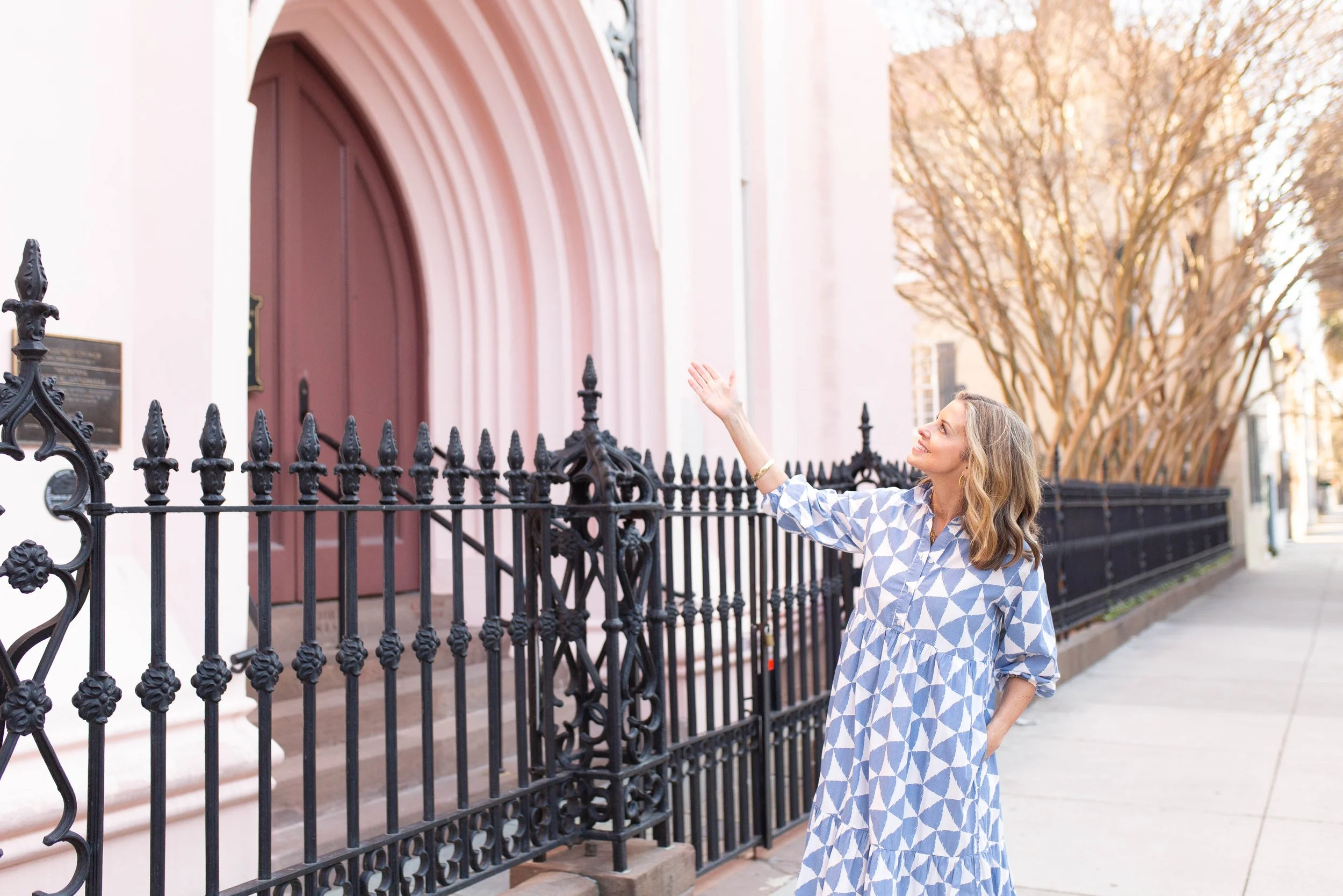 Rachel Fair licensed Charleston tour guide, in a blue and white patterned dress waving outside a pink building with a black wrought iron fence and trees in the background