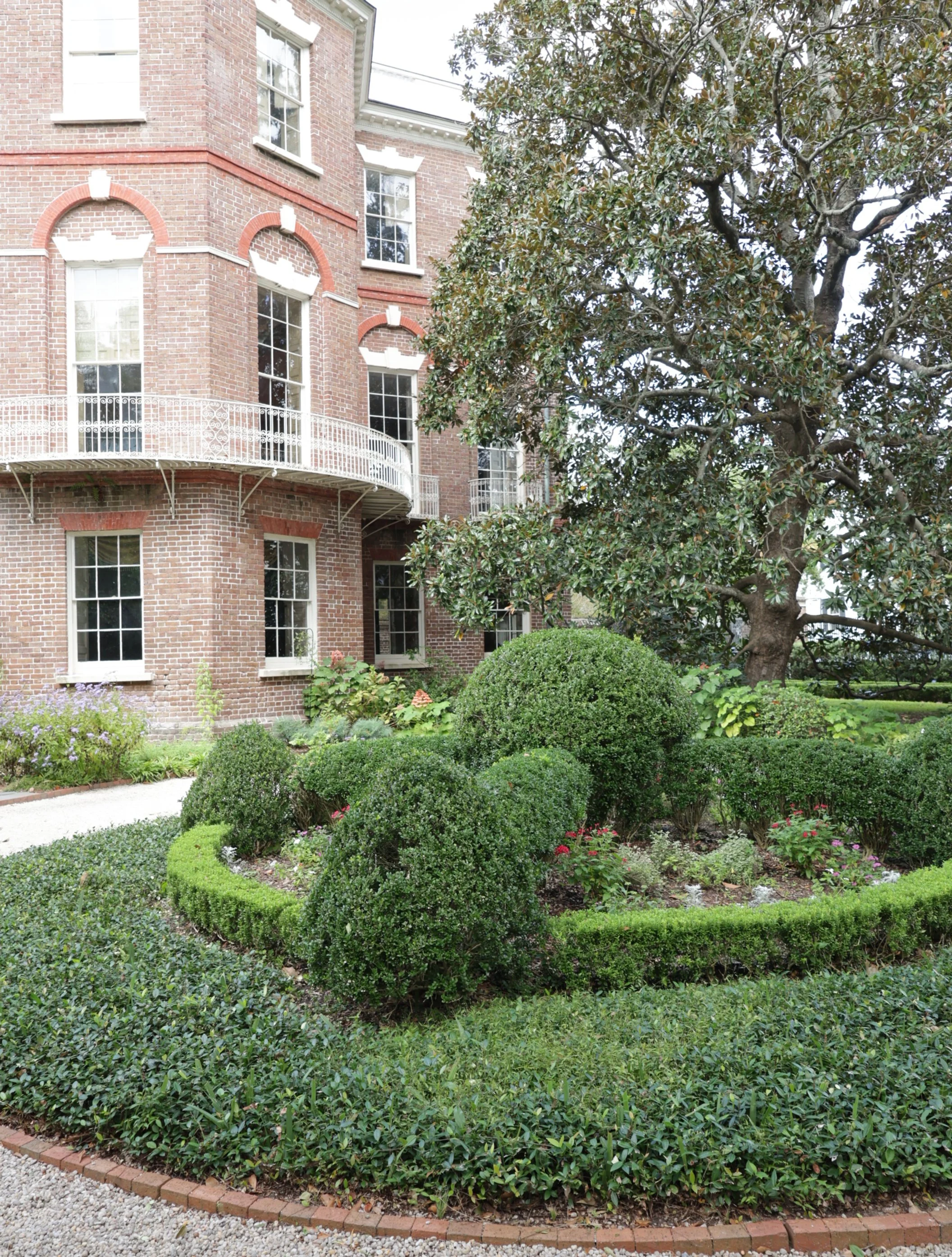 A well-maintained garden with green bushes and flowers in front of a brick building with white-framed windows and a curved balcony.