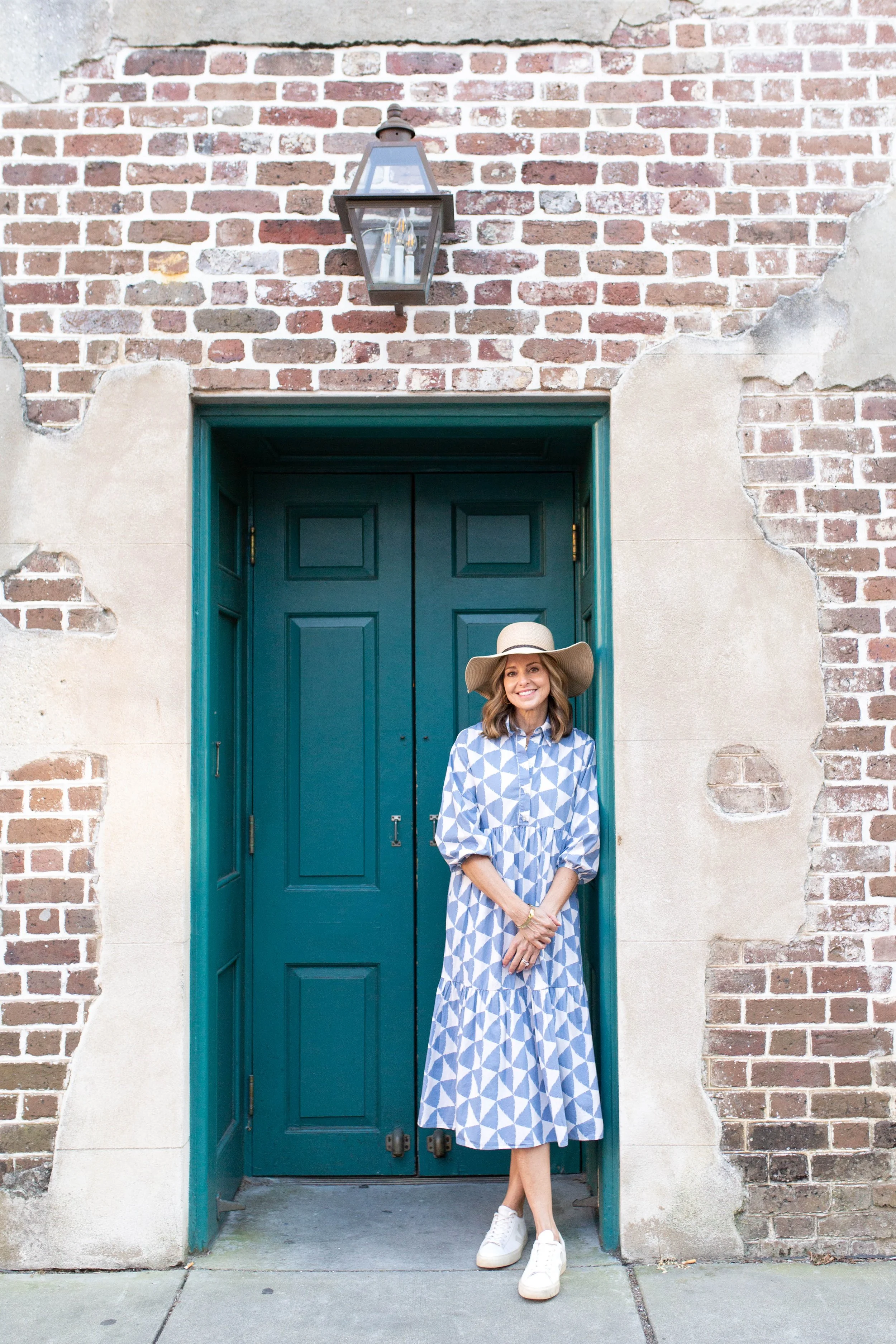 Rachel Fair licensed Charleston tour guide, in a blue and white geometric dress and a wide-brimmed hat standing in front of a dark teal door and an exposed brick wall.