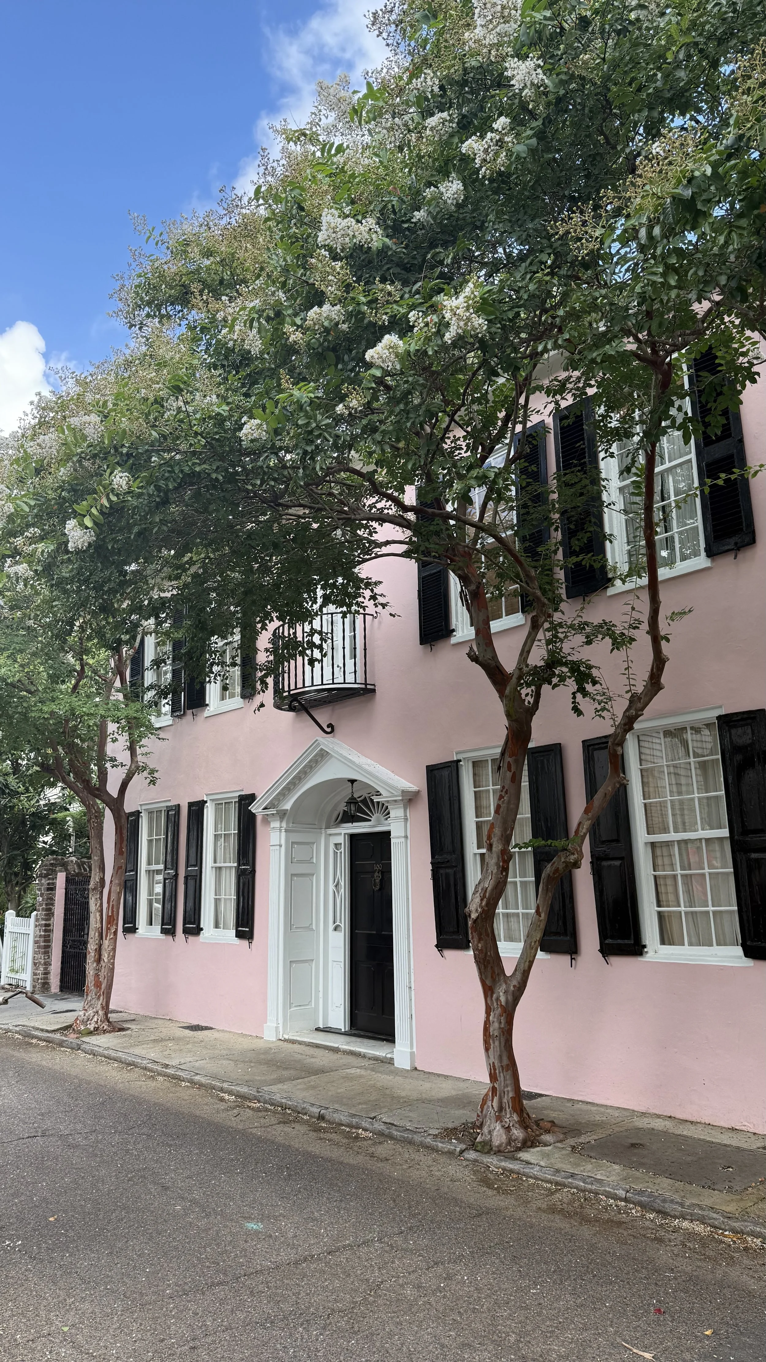 Charleston's iconic pink building with black shutters, white doorframe, and trees visible on the sidewalk under a blue sky with some clouds.