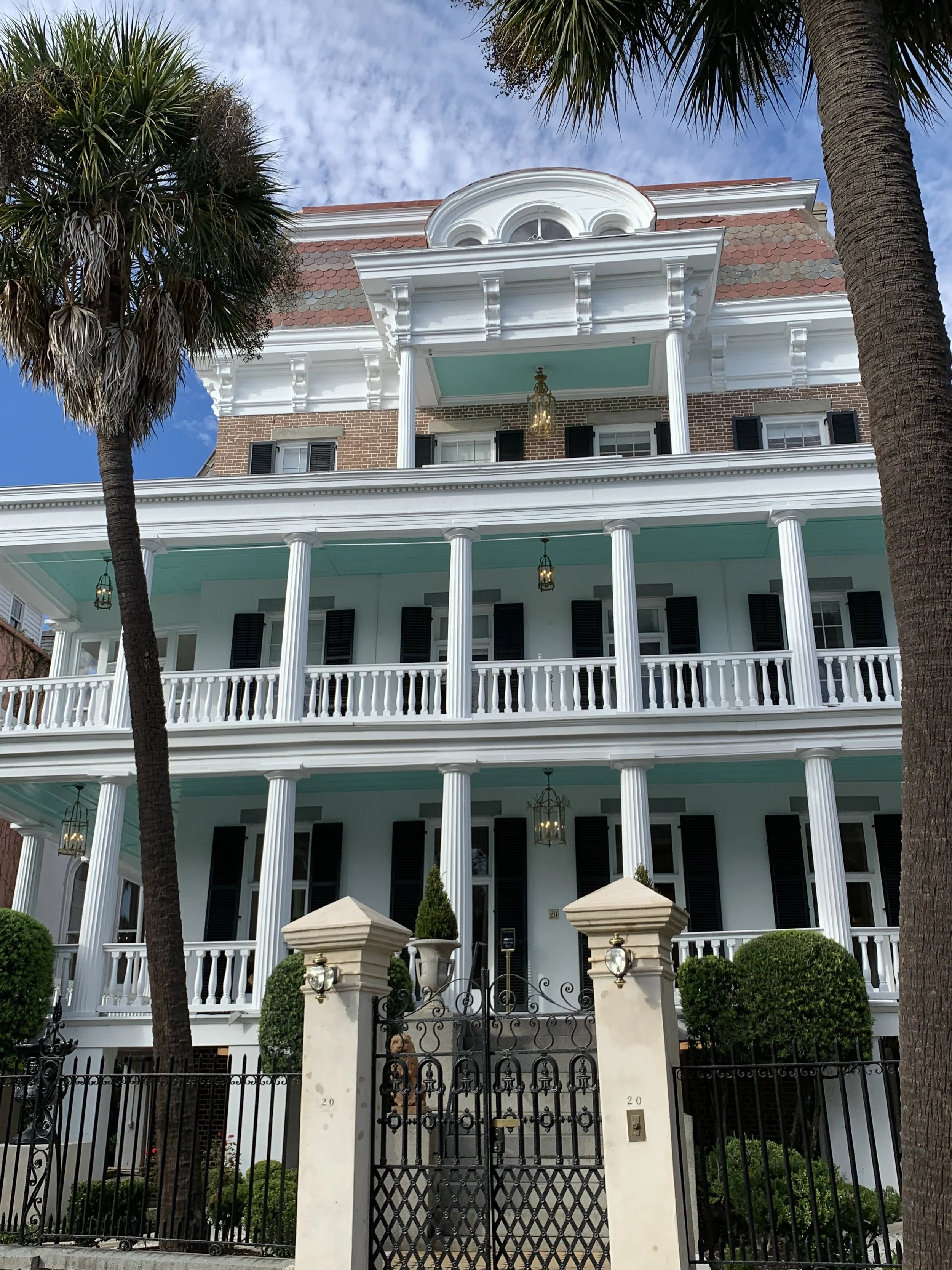 A large white three-story house with a porch, pillars, and black shutters, surrounded by trees and a black iron gate, under a partly cloudy sky.