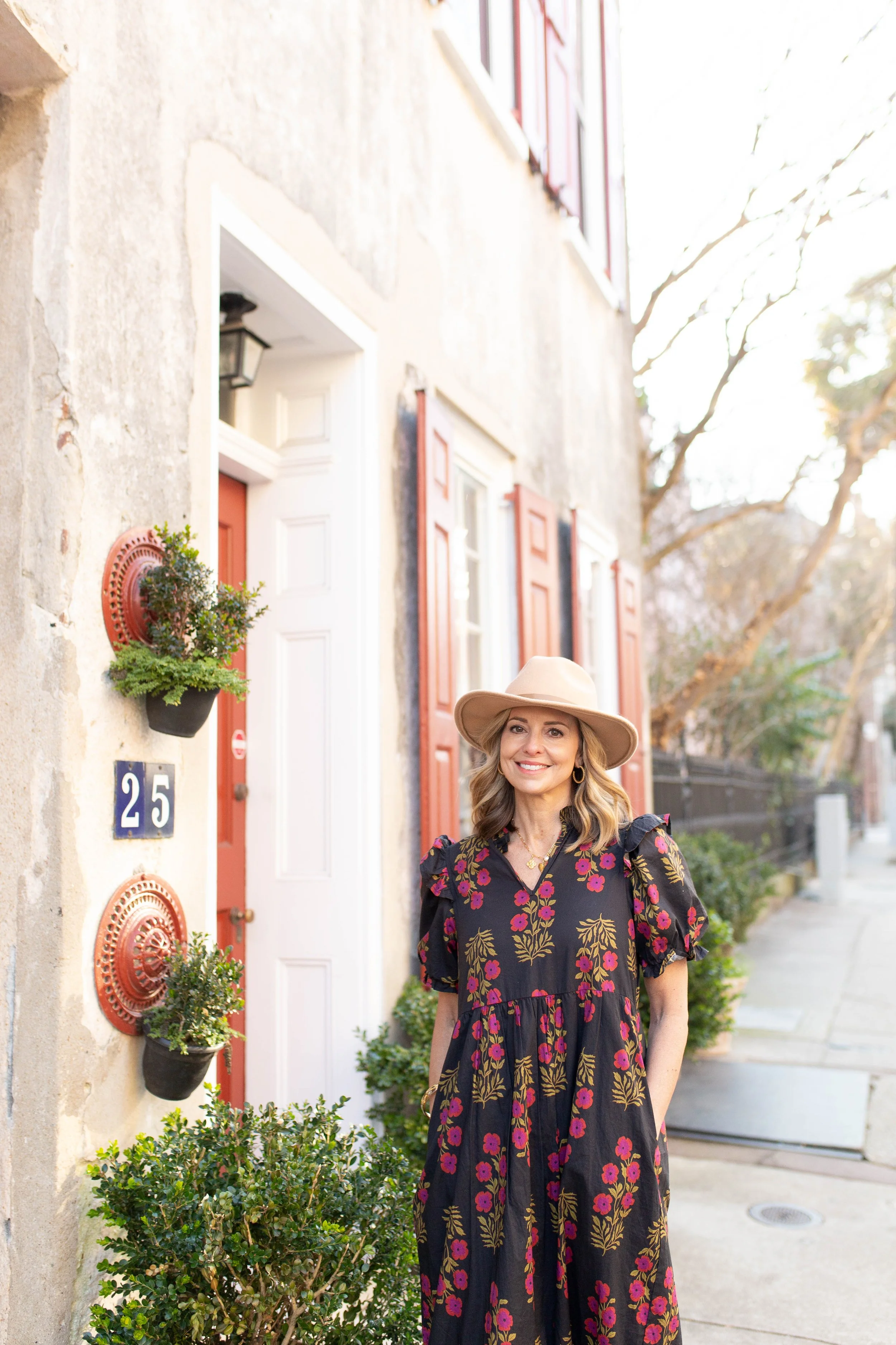 Rachel Fair licensed Charleston tour guide, standing on a sidewalk outside a house wearing a floral dress and a wide-brimmed hat, smiling at the camera.