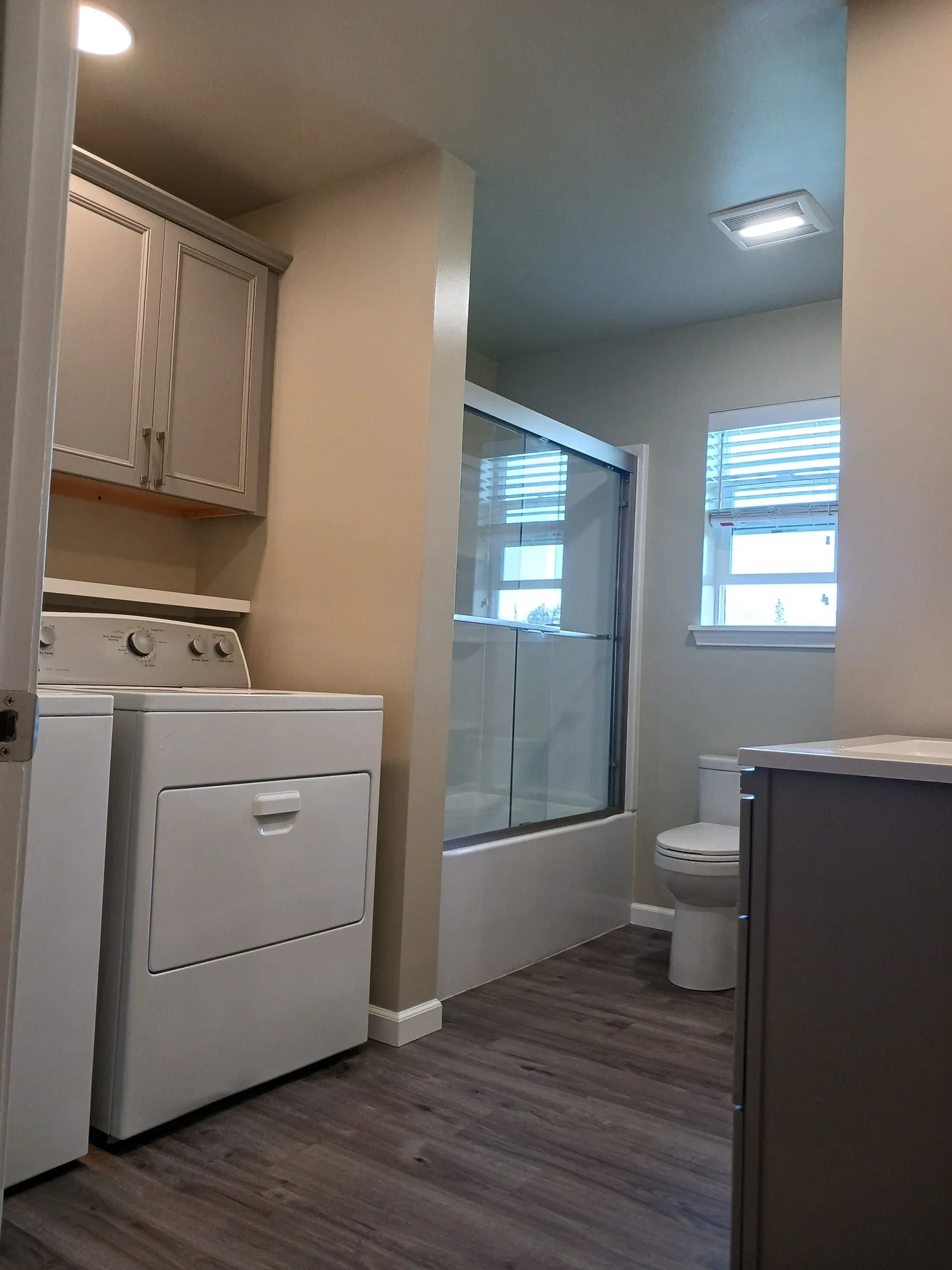 Inside a bathroom with a laundry area, featuring a washing machine, a toilet, a shower with a glass door, and a small window with blinds.