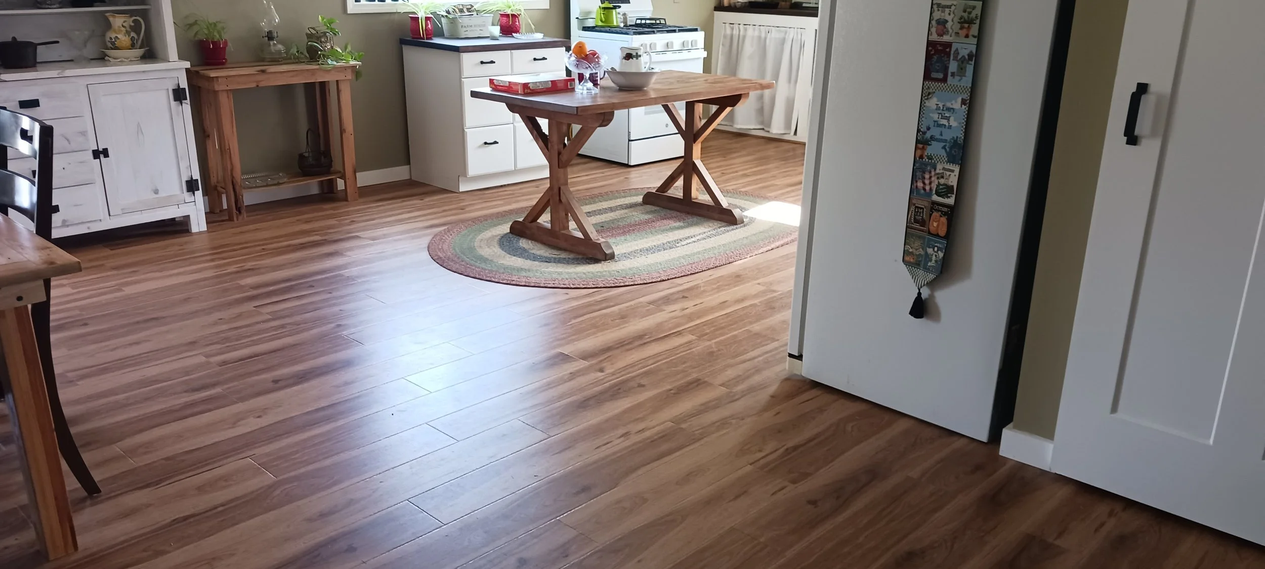 Kitchen with wood flooring, white cabinets, a small table with a plaid tablecloth, and a round multicolored rug. Countertop appliances and plants are visible near the window.