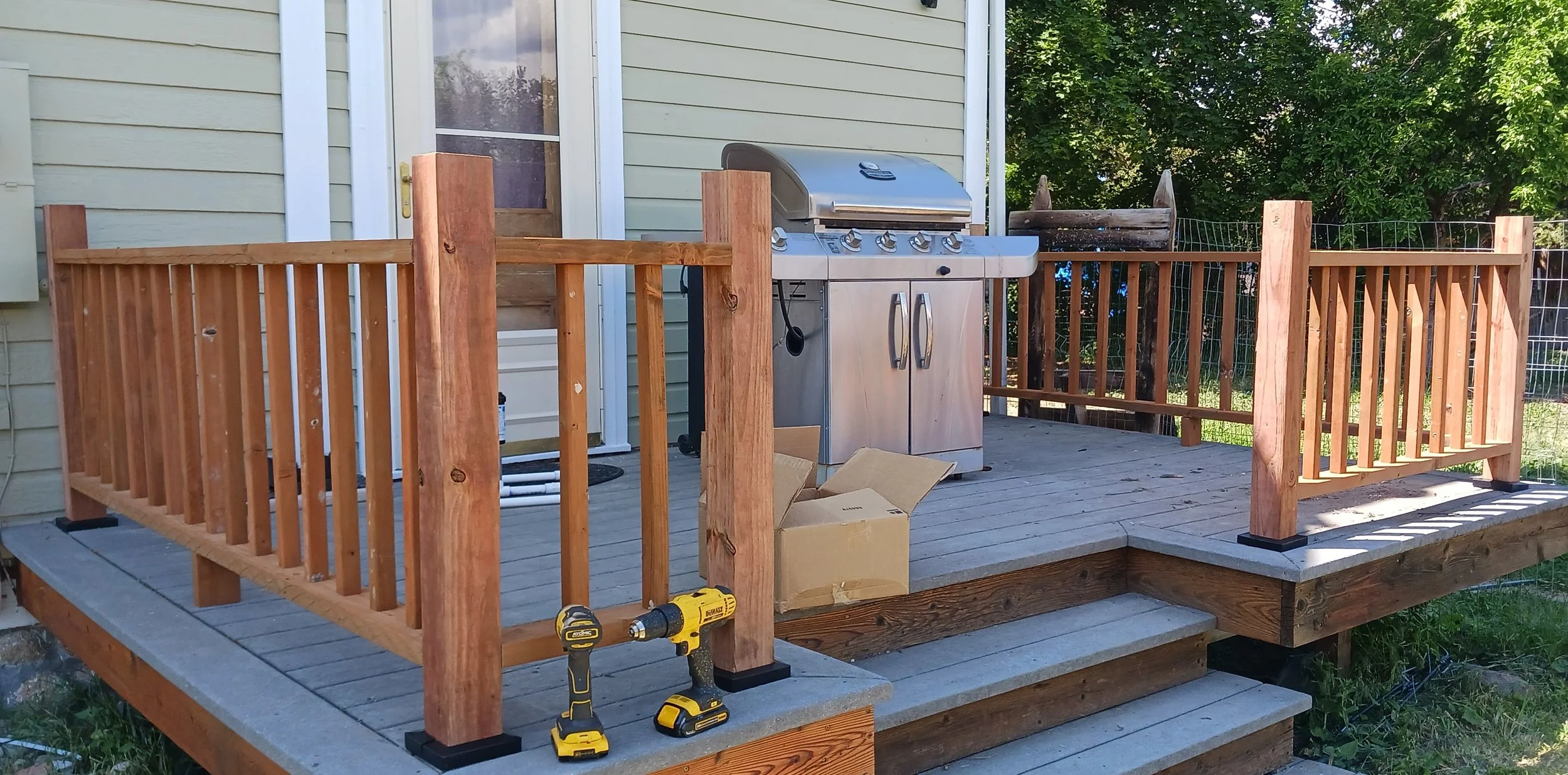 A newly built wooden deck attached to a house, with a stainless steel grill and tools placed on it, and a cardboard box on the floor. The deck is surrounded by a wooden railing and overlooks a backyard with green trees.