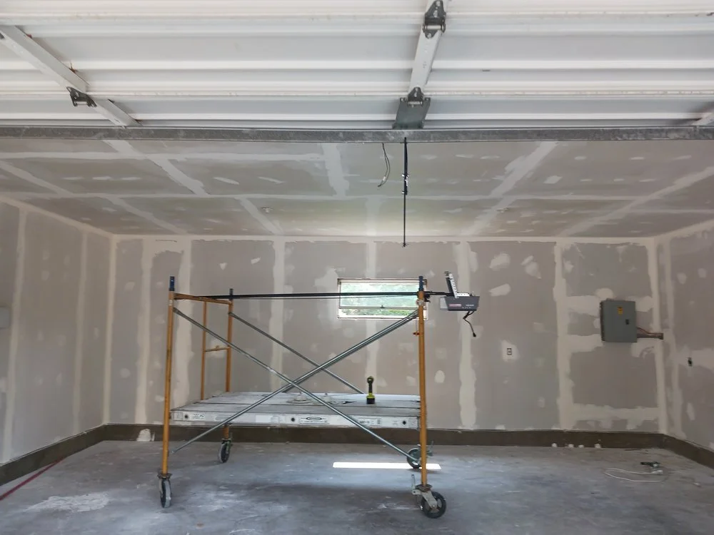 Empty room with drywall walls, a small window, and a ceiling wired for lighting. There is a scaffolding platform with tools on it, and some construction materials on the floor.