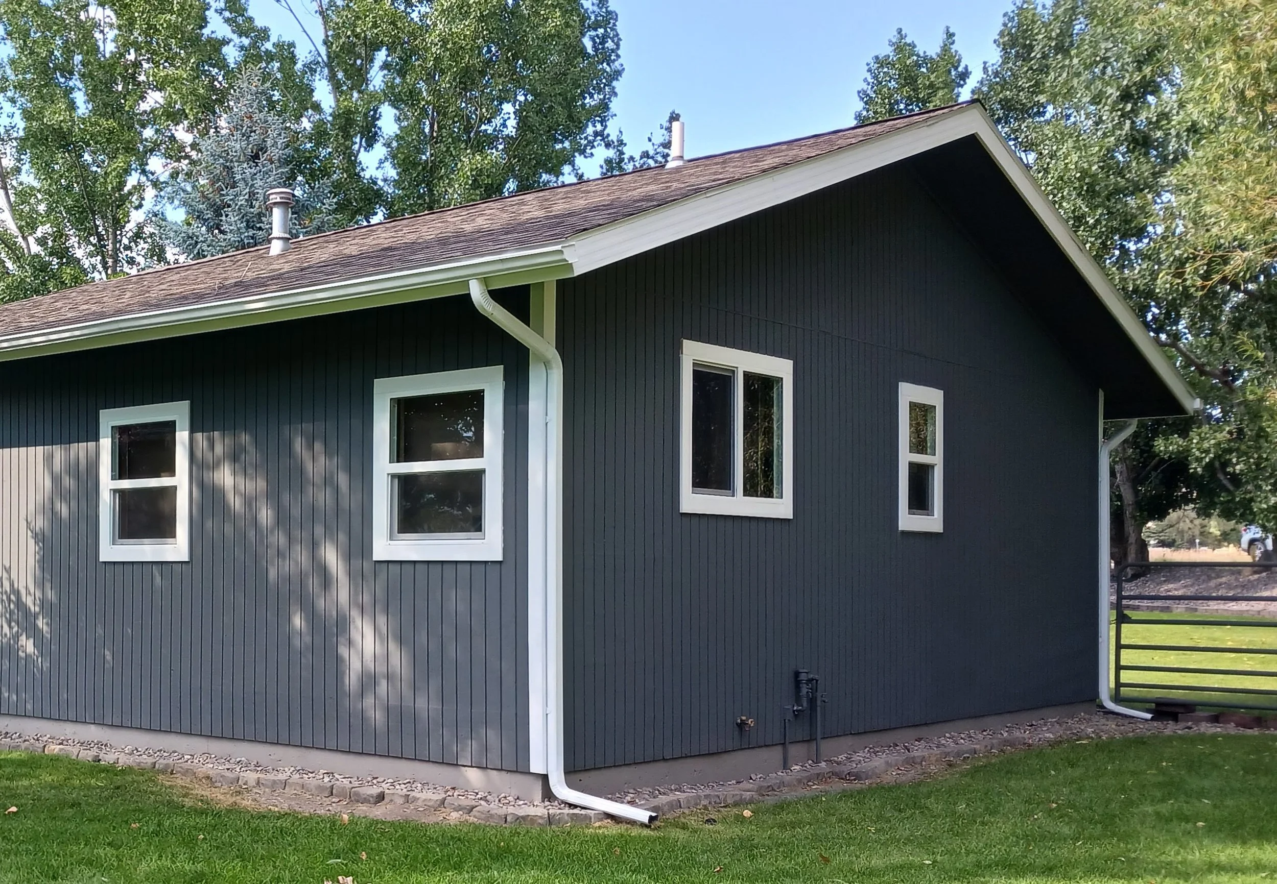 Side view of a house with black vertical siding and white trim, three white-framed windows, a shingled roof, and a small grassy yard.