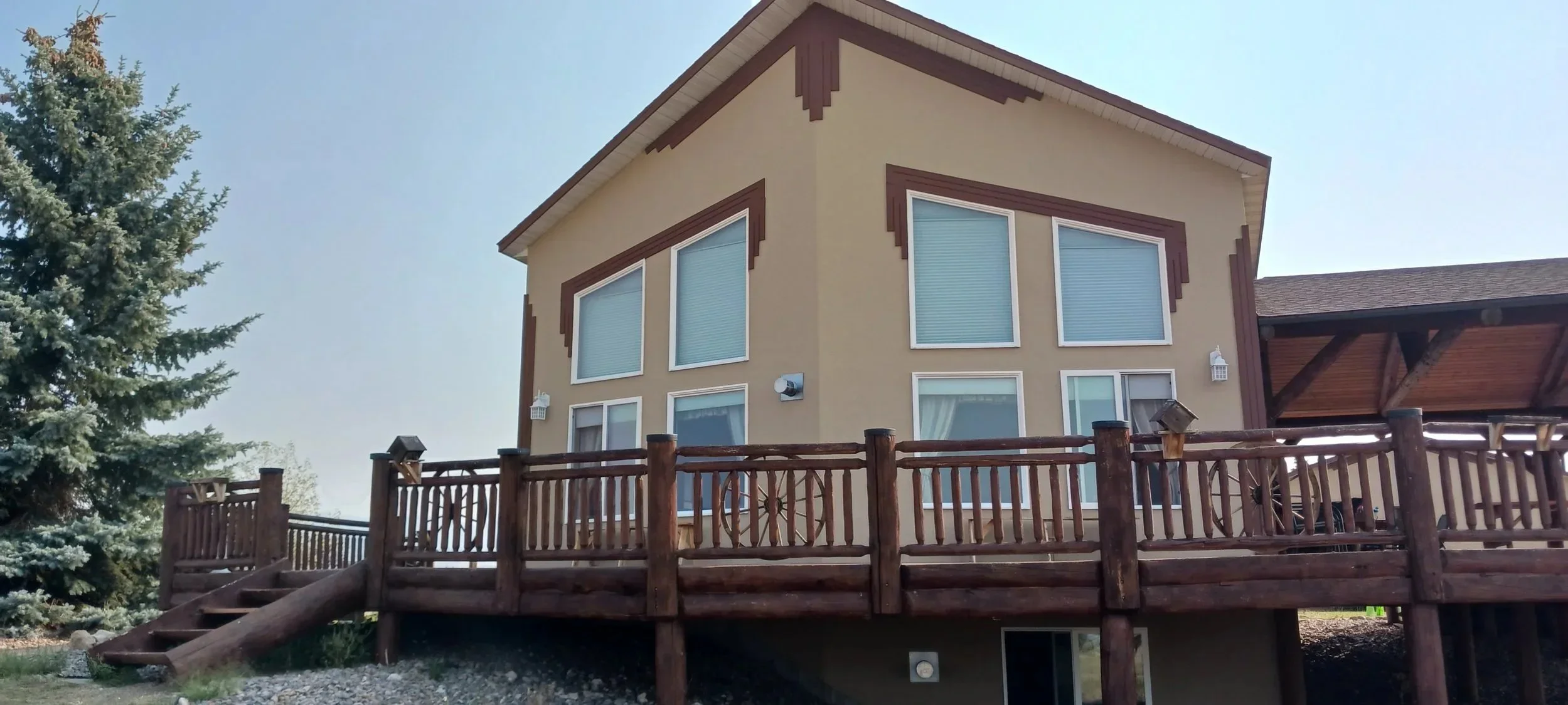 A two-story house with beige walls and brown trim, large windows with blinds, and a wooden deck with stairs and a railing, surrounded by trees and a clear sky.