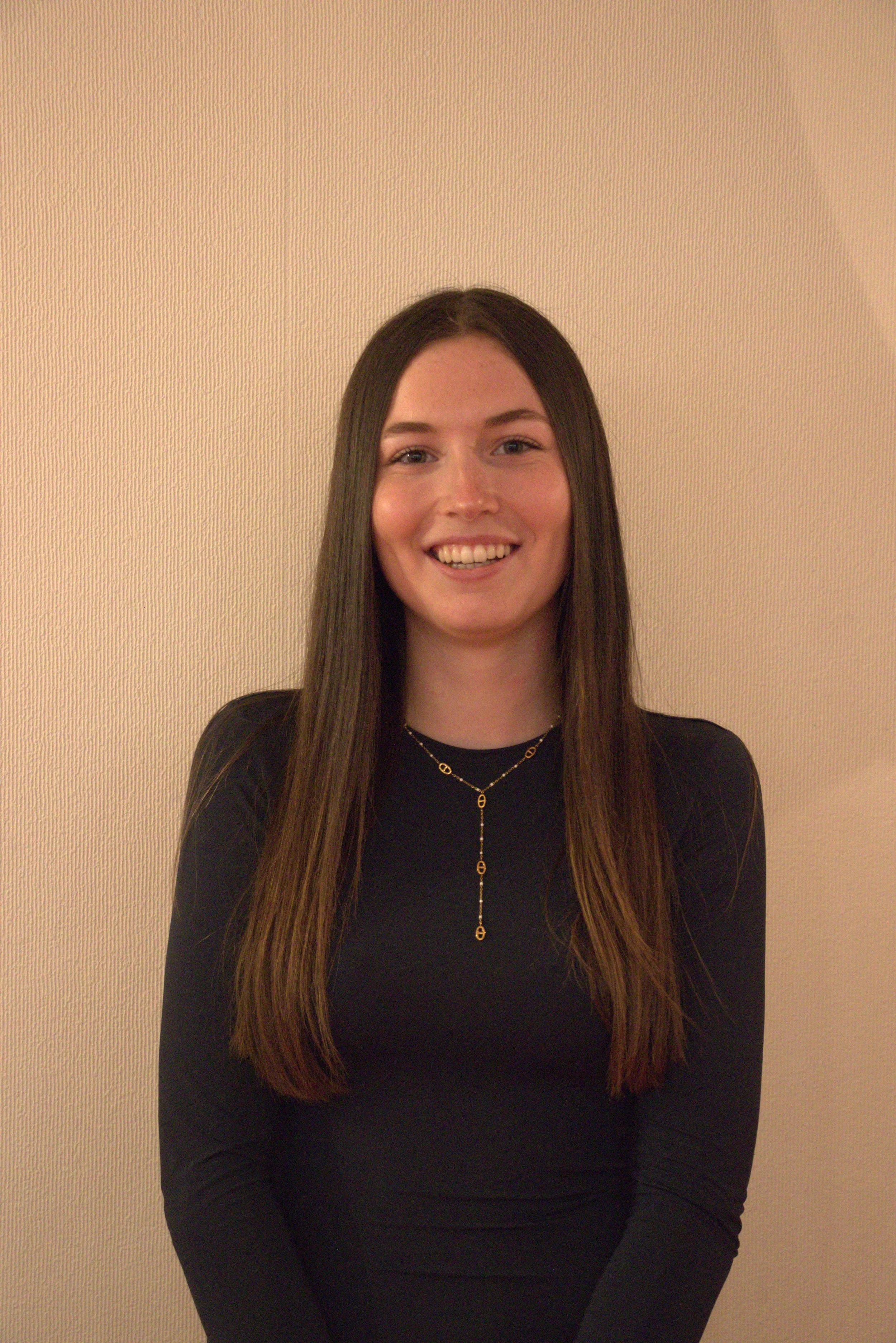 Une femme souriante avec de longs cheveux bruns portant une robe noire et un collier doré, posant contre un mur beige.