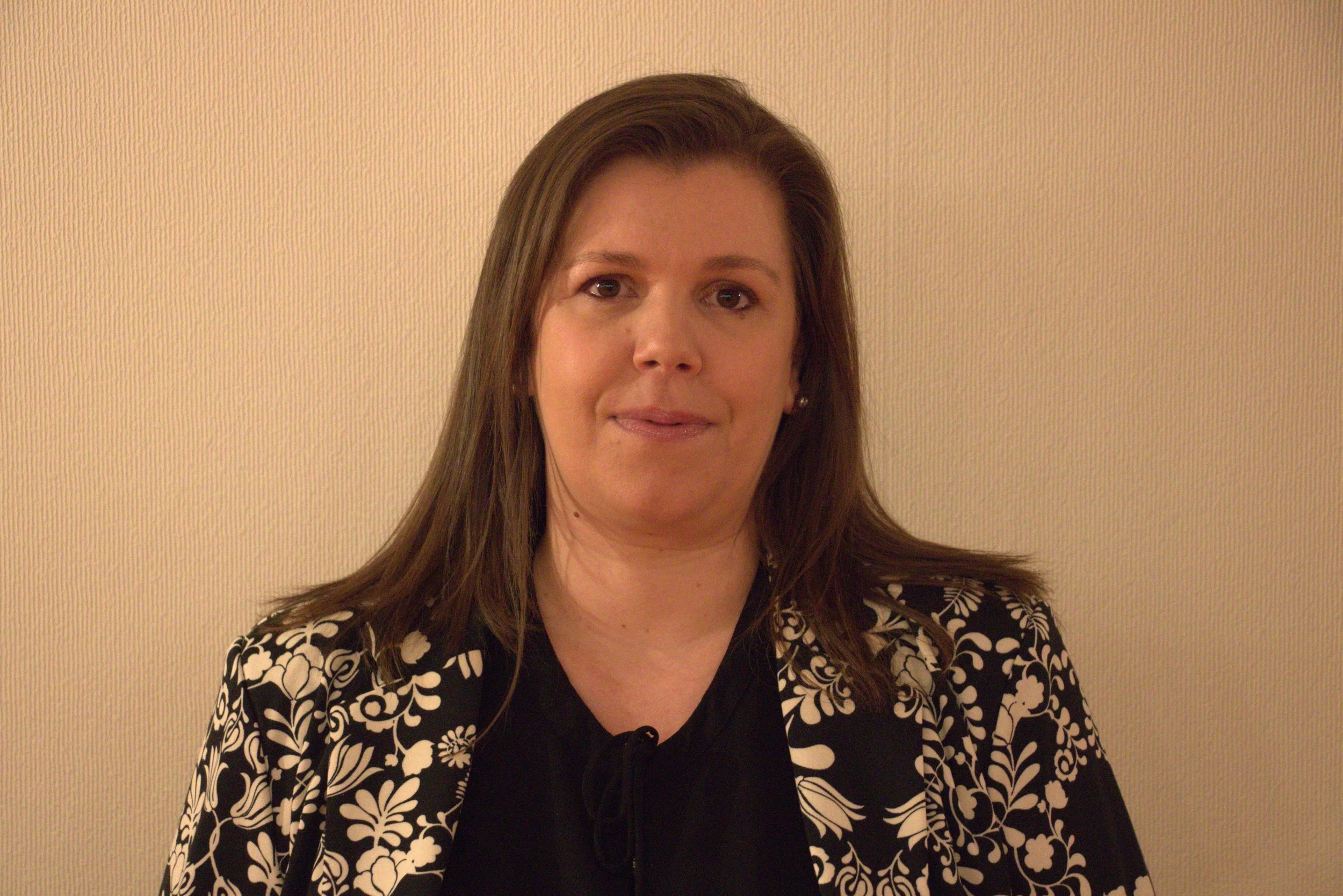 Une femme avec des cheveux bruns, portant une veste noire à motifs blancs, se tenant devant un mur beige.