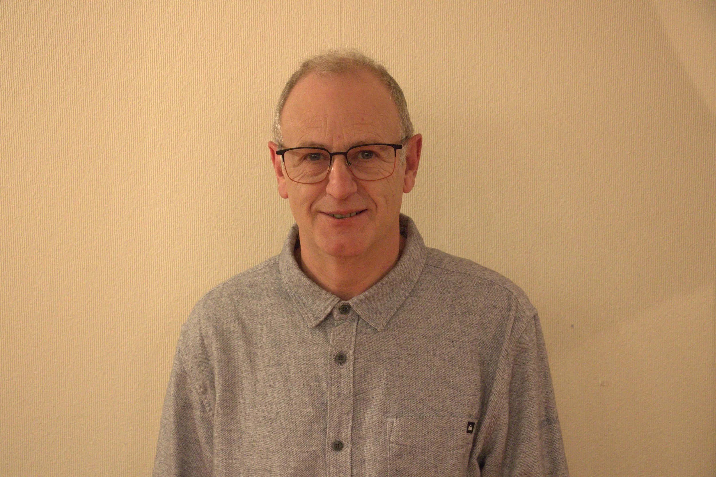 Un homme moustachu, portant des lunettes, un pull gris en regardant l'objectif, contre un mur beige.