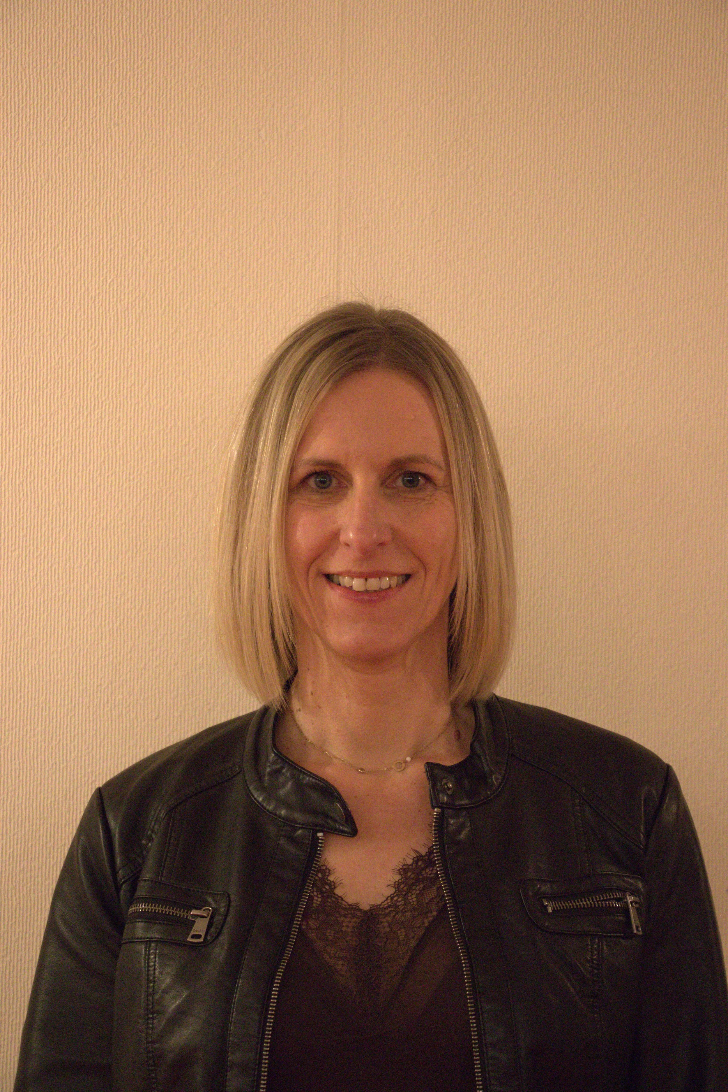 Une femme aux cheveux blonds, portant une veste en cuir noir, souriante, devant un mur beige.