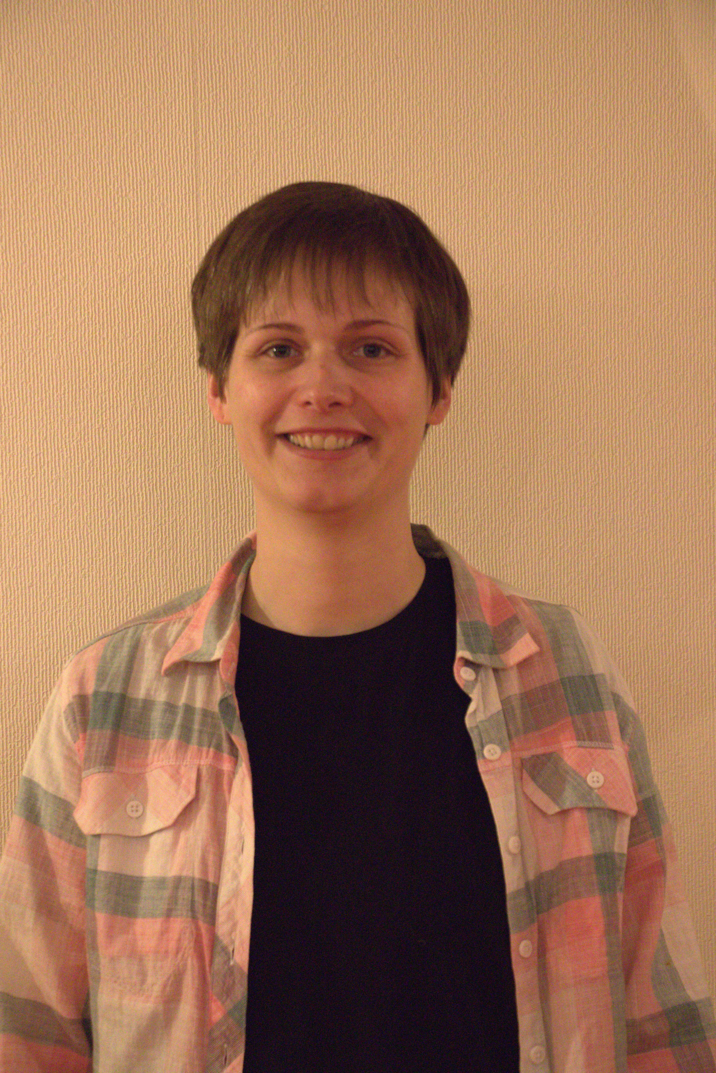 Jeune femme souriante avec une chemise à carreaux rose et verte, en arrière-plan un mur beige.