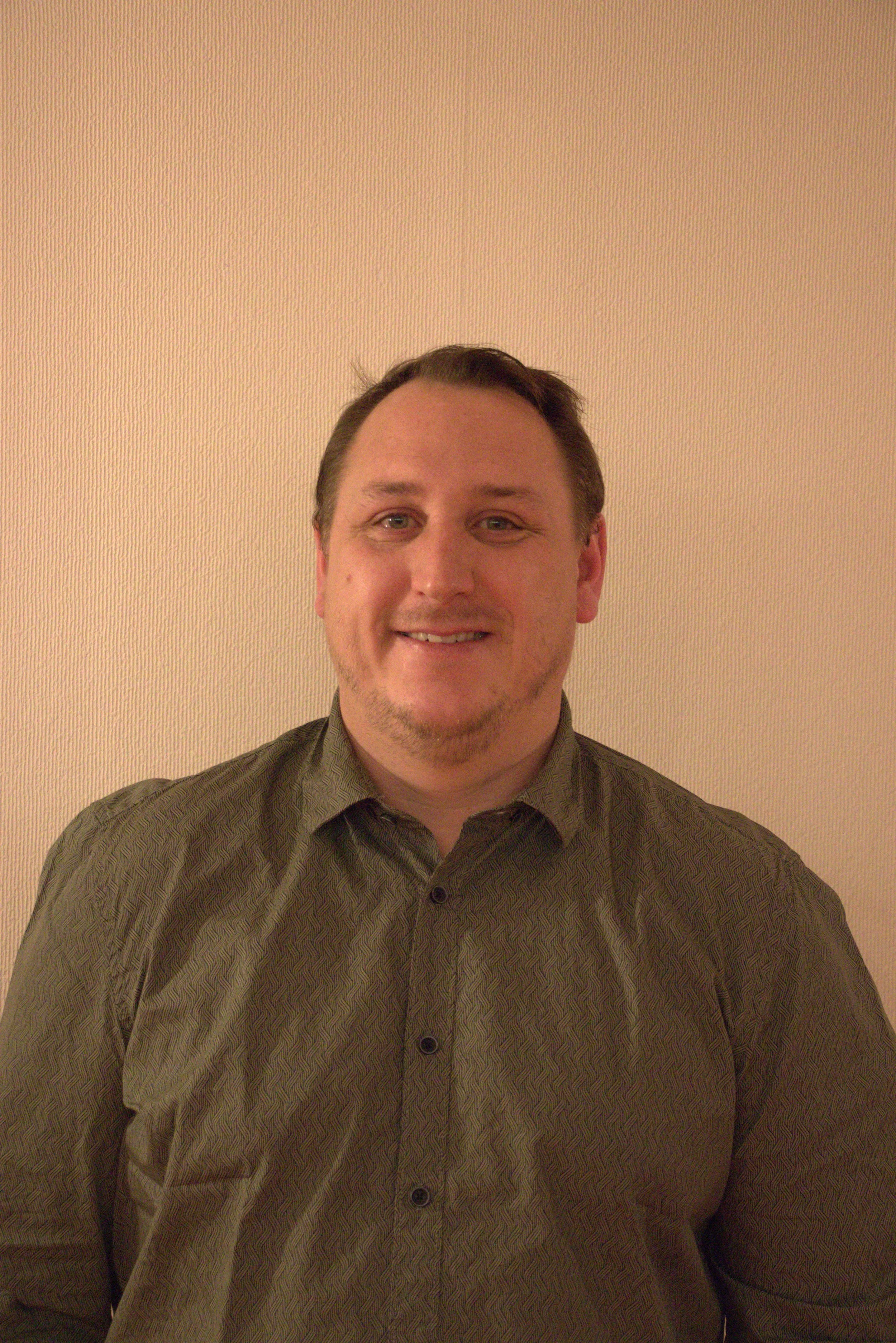 Un homme souriant portant une chemise à motifs, posé devant un mur beige.