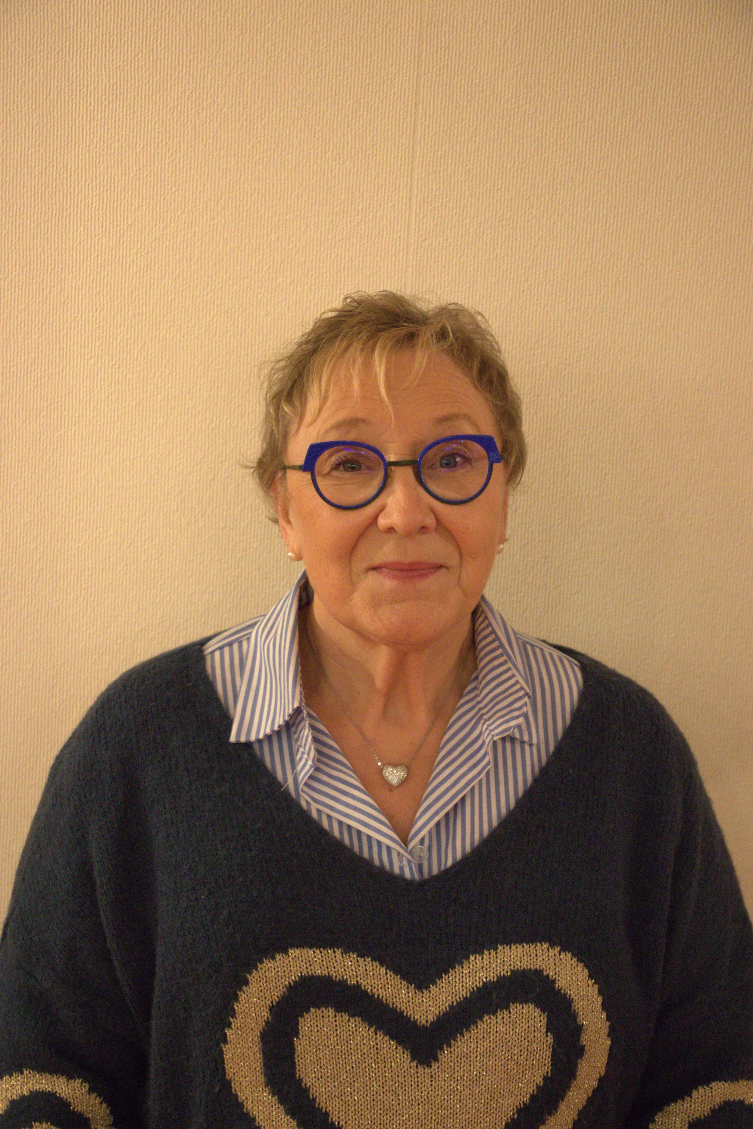 Une femme âgée portant des lunettes rondes bleues, une chemise à rayures et un pull noir avec un motif de cœur beige, posant devant un mur beige clair.