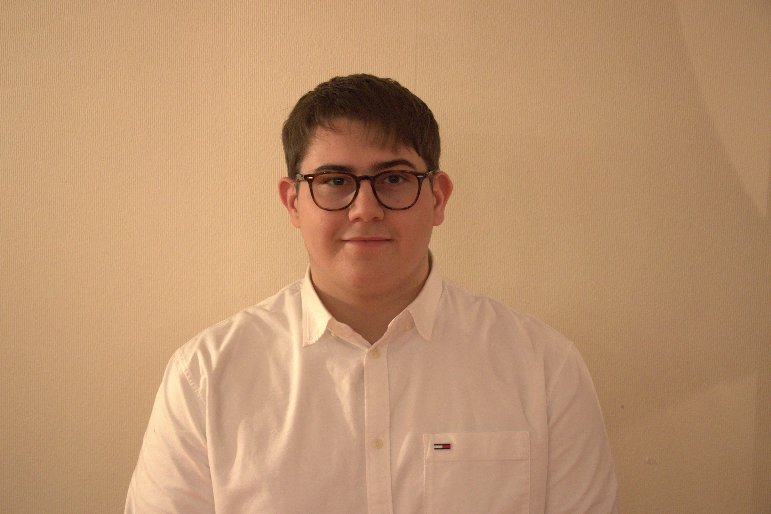 Jeune homme portant des lunettes et une chemise blanche, posant devant un mur beige.