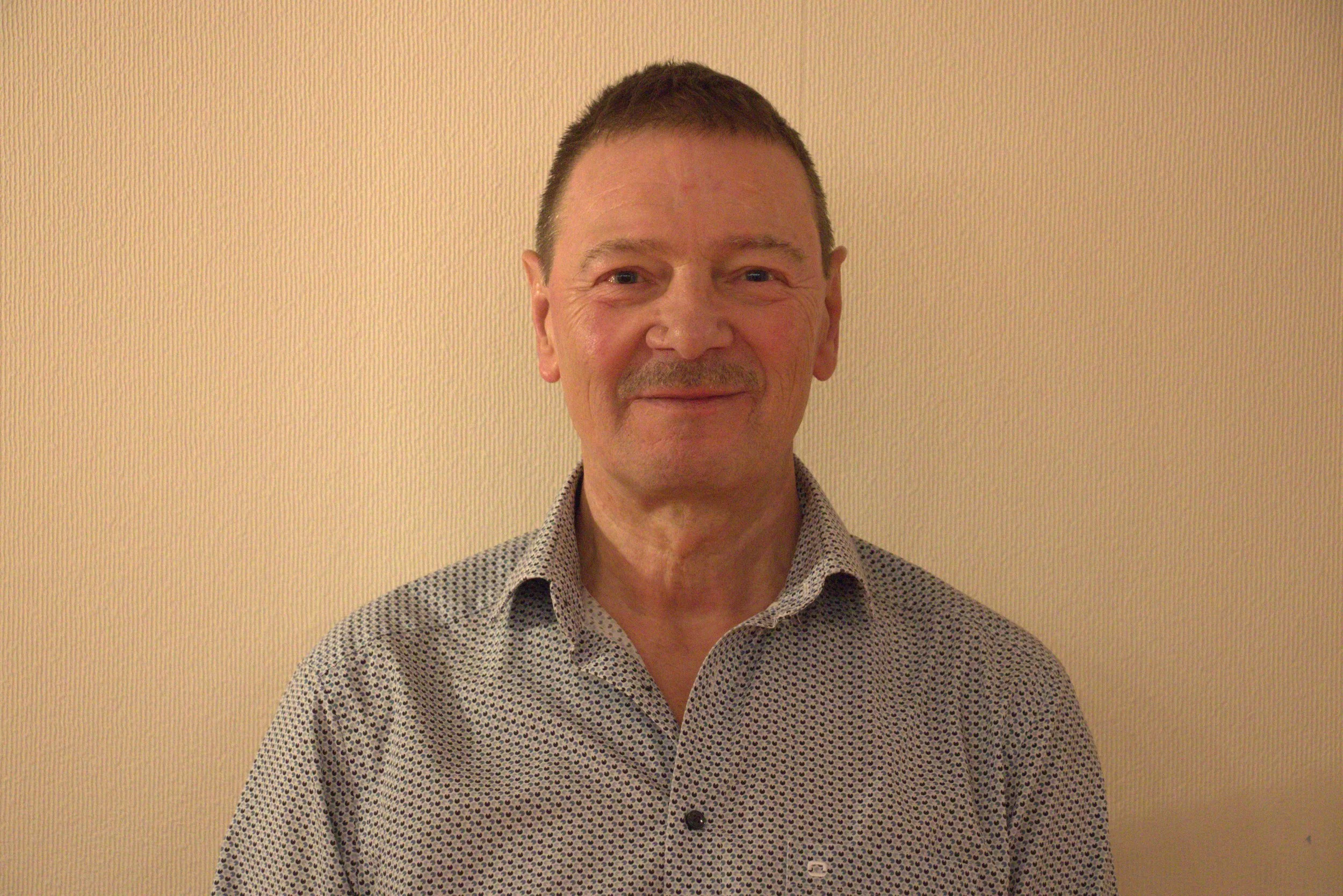 Un homme souriant avec des cheveux courts et une chemise à motifs posant devant un mur beige.