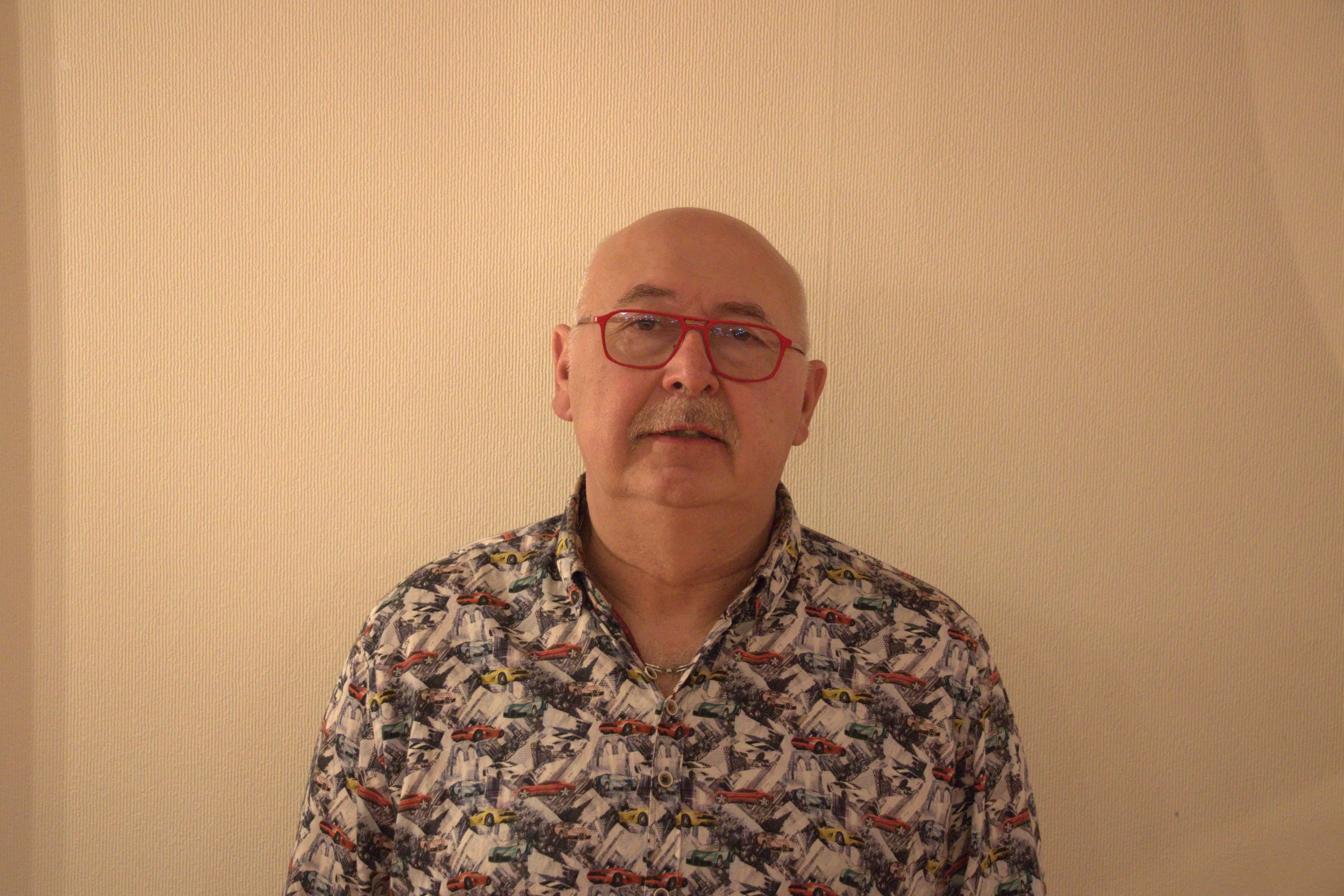 Un homme d'âge moyen portant des lunettes rouges, une chemise à motif de voitures, se tient devant un mur beige.