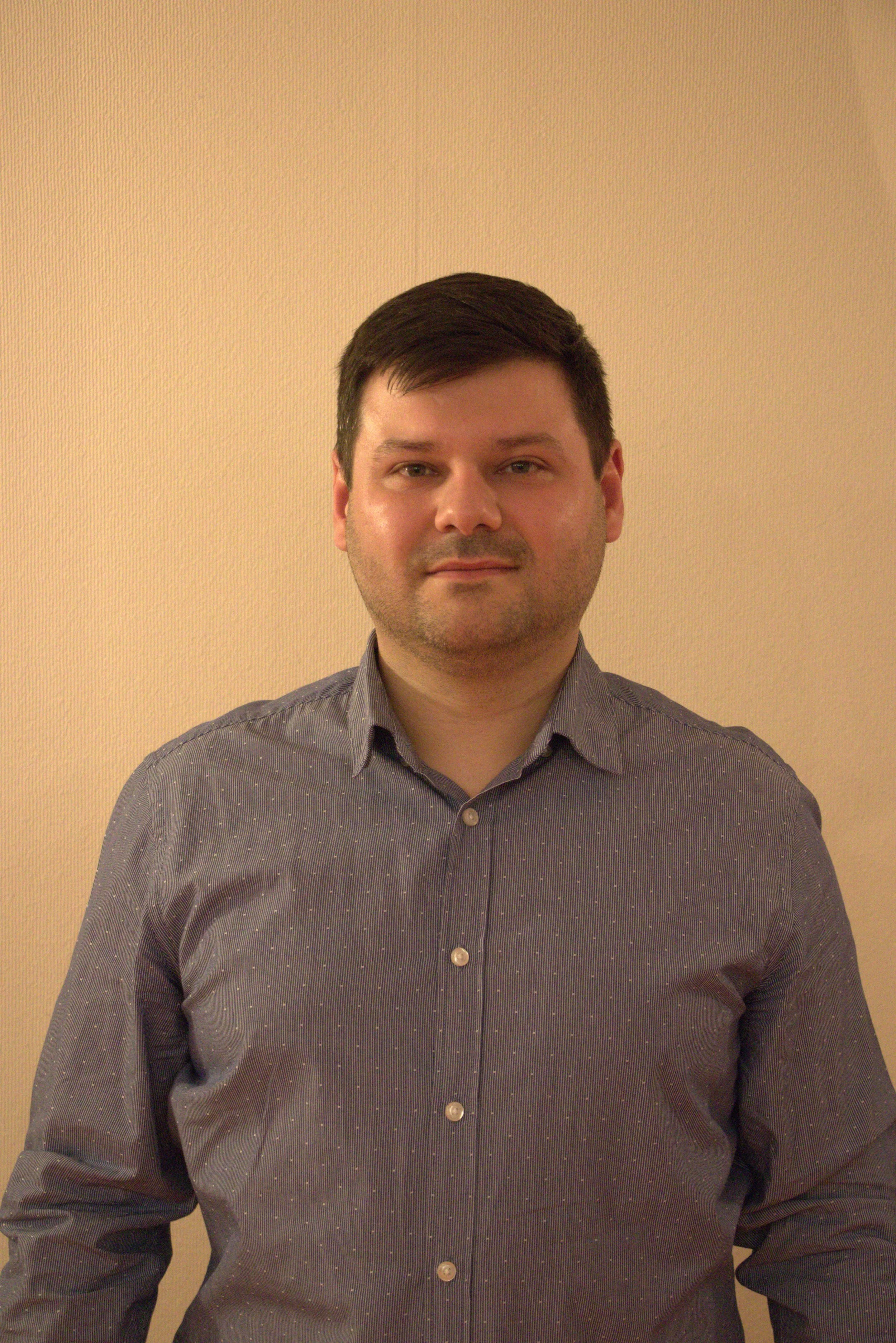 Portrait d'un homme portant une chemise gris foncé à petits pois, debout devant un mur beige.