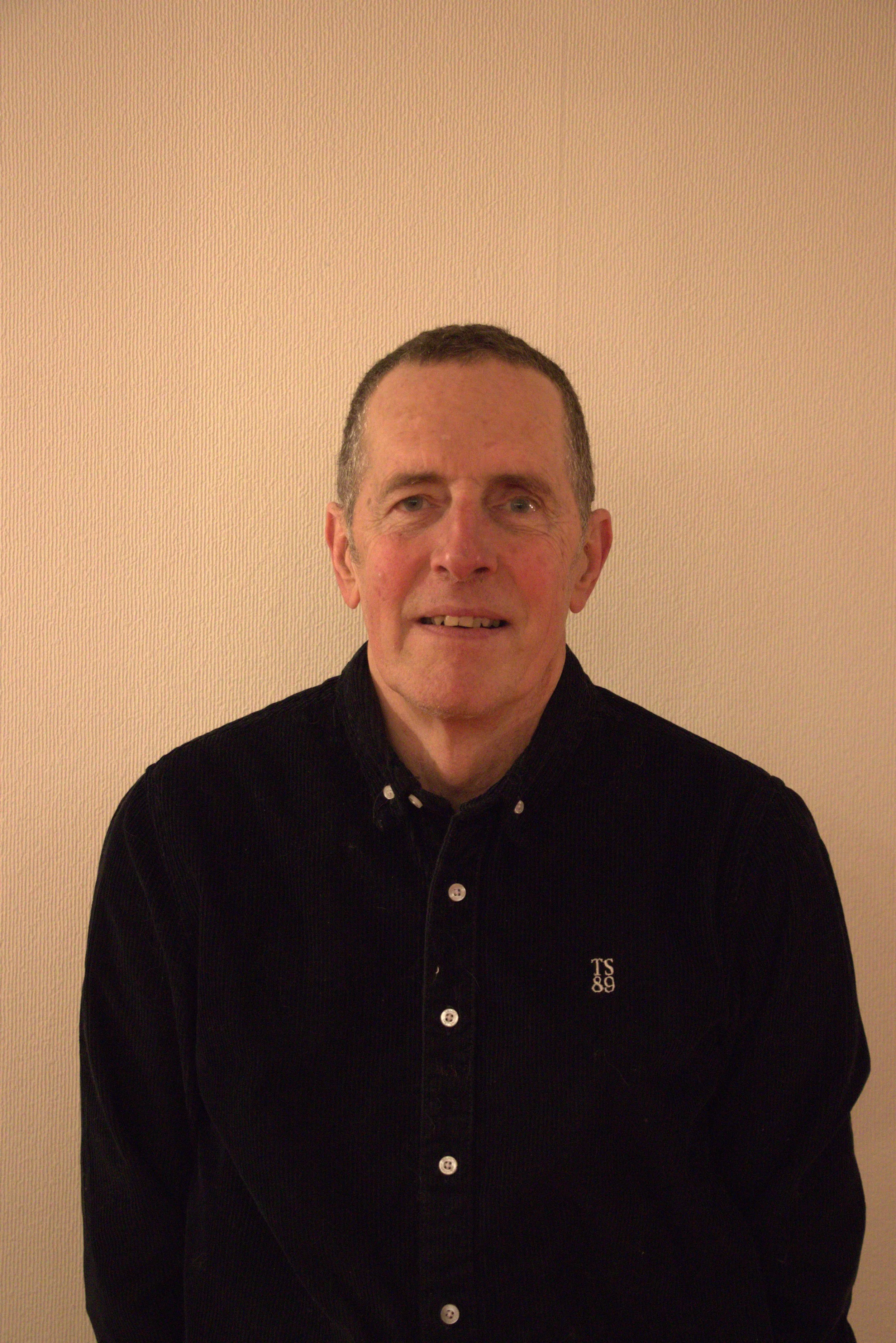 Portrait d'un homme avec des cheveux courts, portant une chemise noire en velours côtelé, devant un mur beige