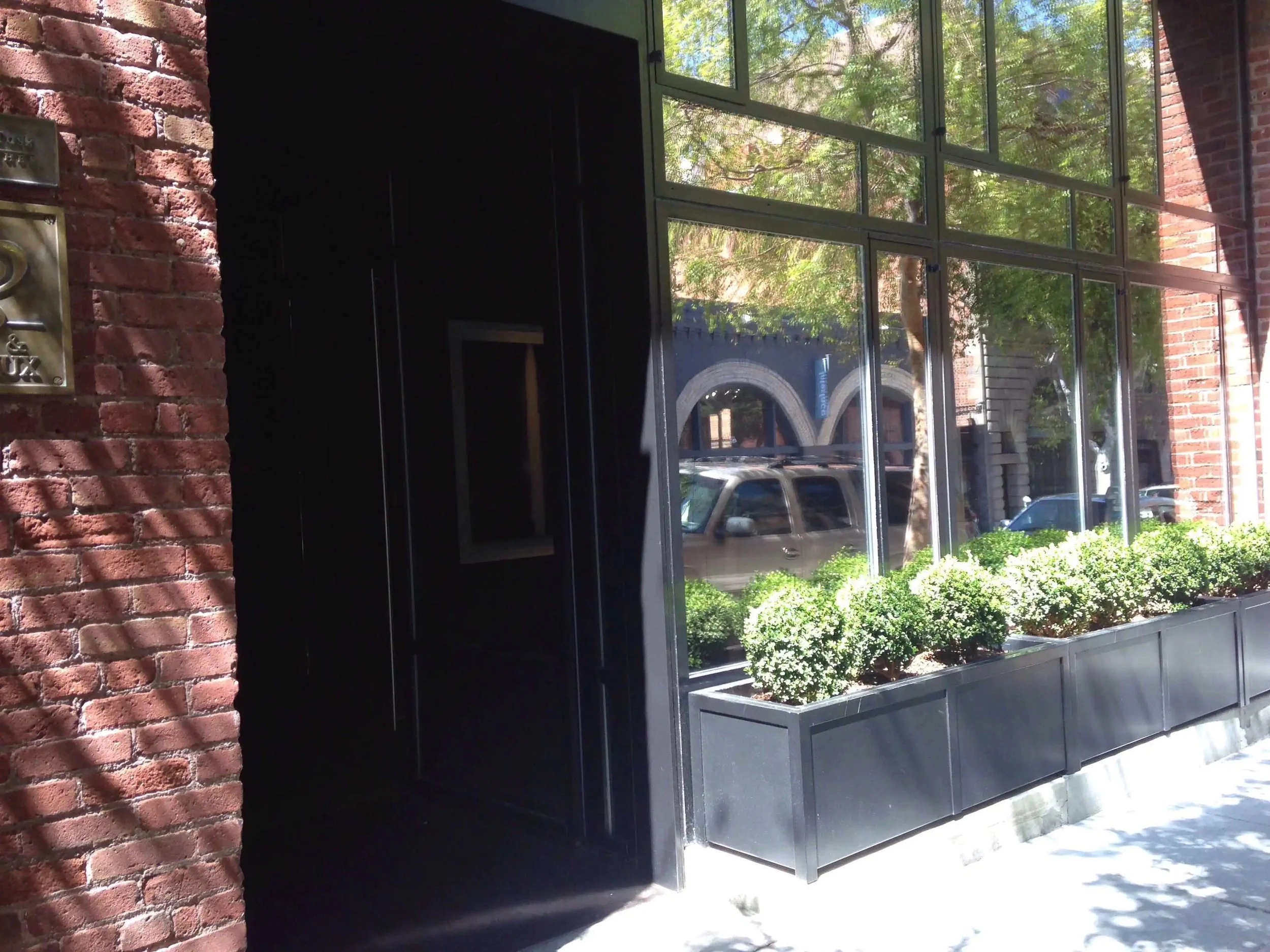 A building with a large glass window and brick wall, with plants in black planter boxes outside, and a street with parked cars and trees visible through the window.
