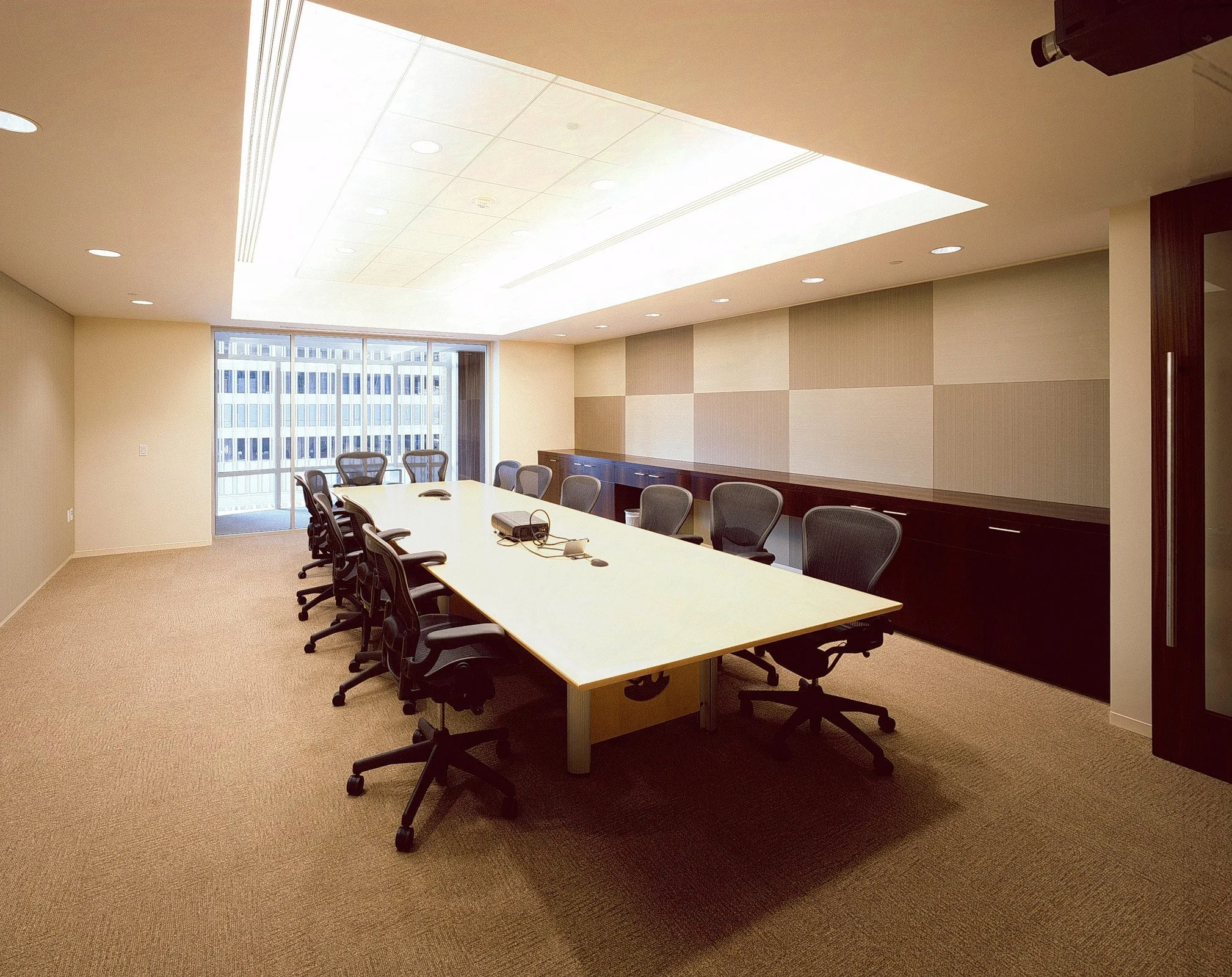Empty conference room with a long table, ten black office chairs, and a large window showing a city building outside.